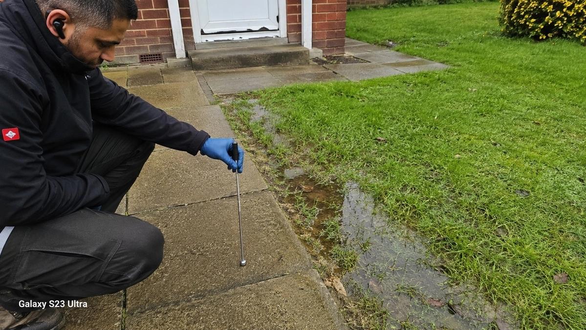 Engineer holding an acoustic listening stick against a driveway slab beside a lush, waterlogged lawn patch.