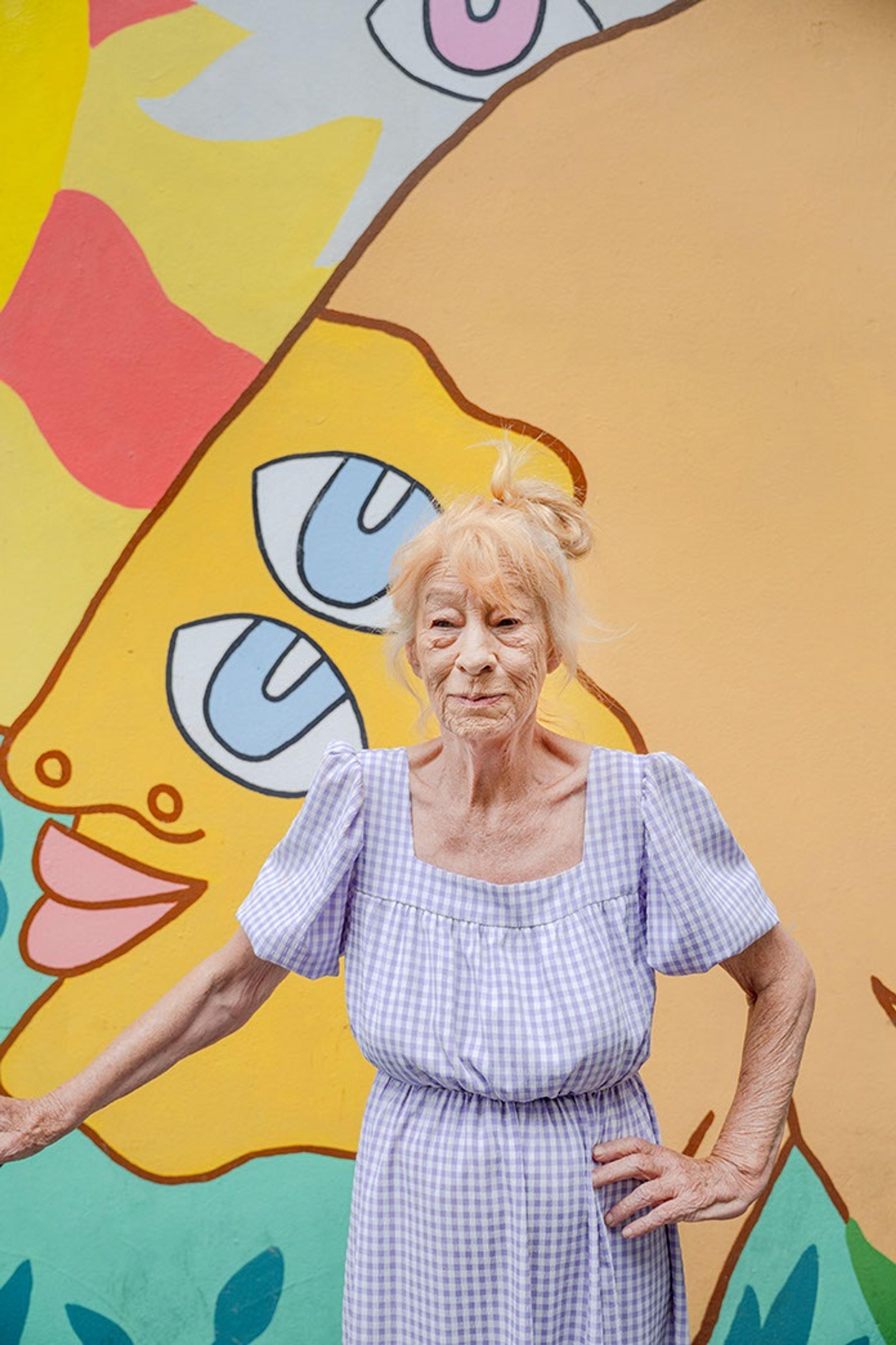 a woman with her hand on her hip stands facing the camera with her hand on her hip, in front of a bright mural