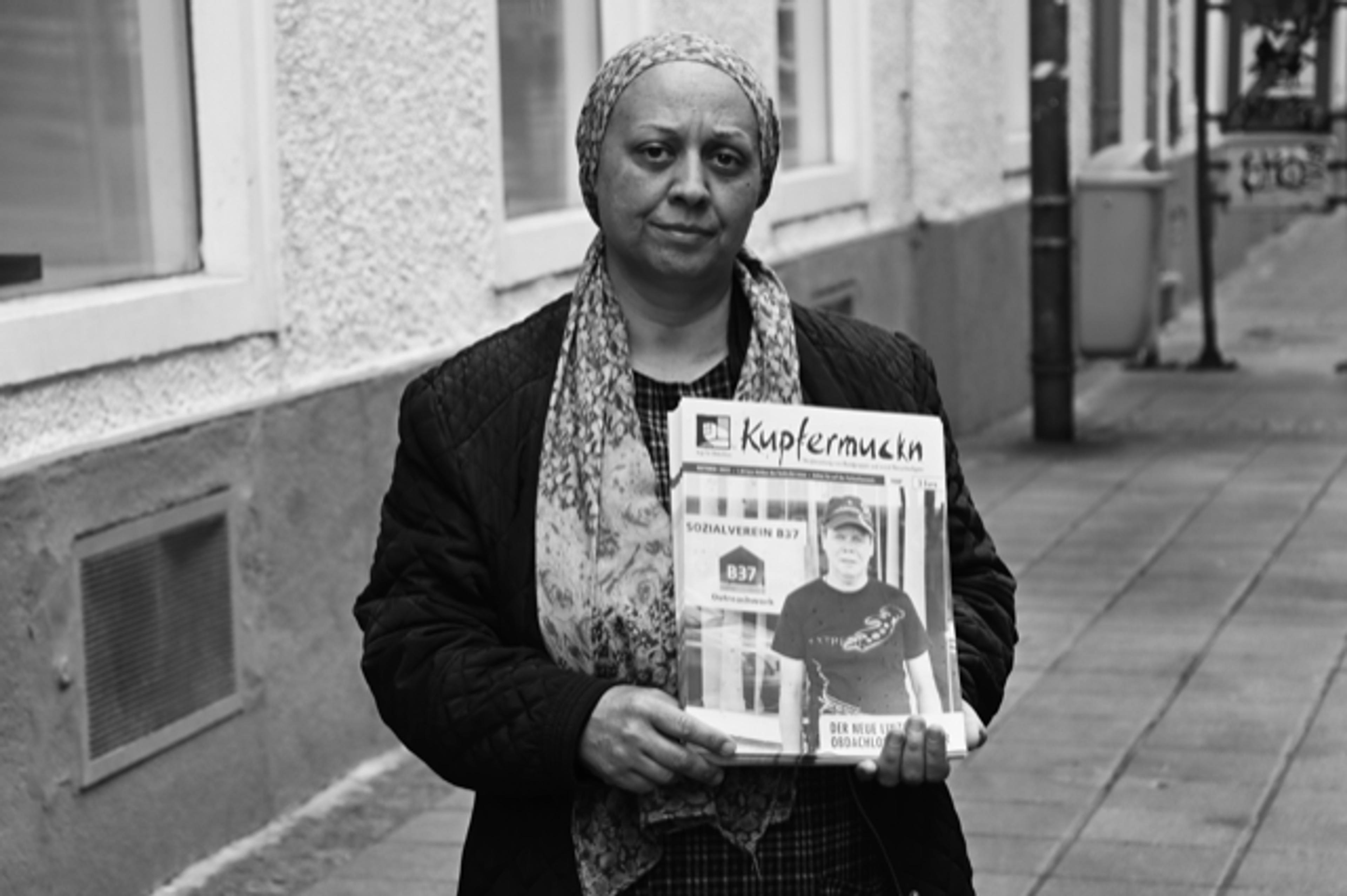 Kupfermuckn vendor Ramona stands with a bundle of magazines in a head scarf. The photo is in black and white.