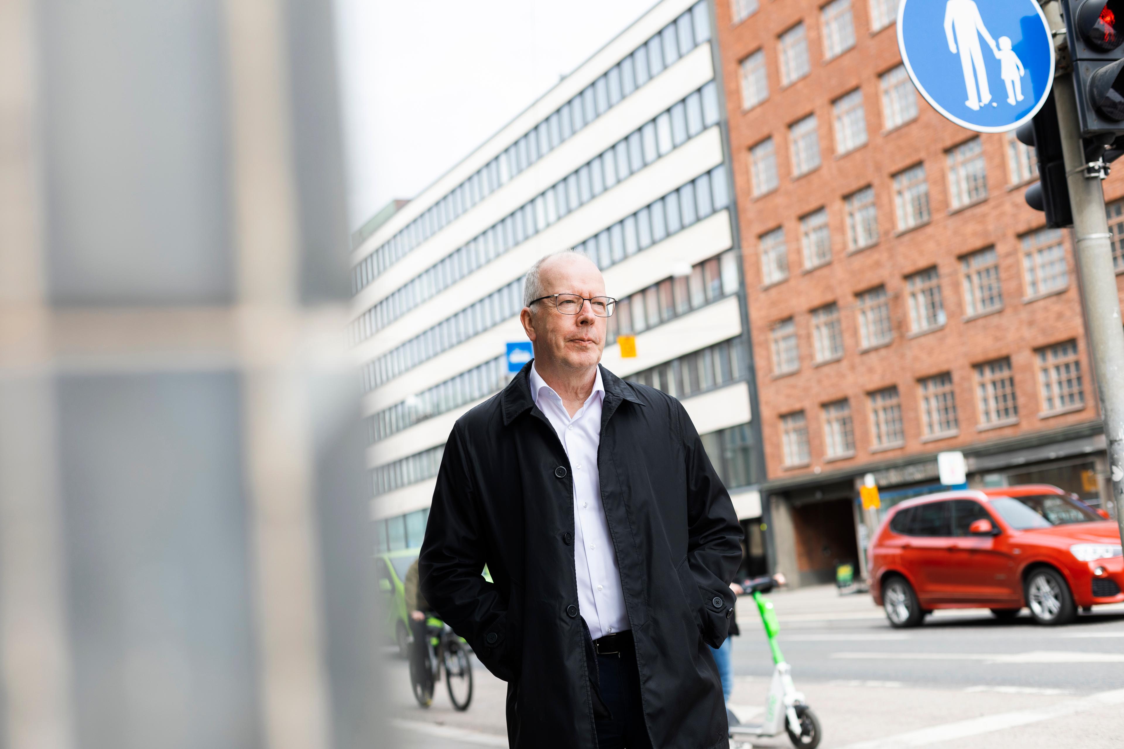 Juha Kaakinen wears glasses a white shirt and black jacket. He is photographed walking down a busy street.