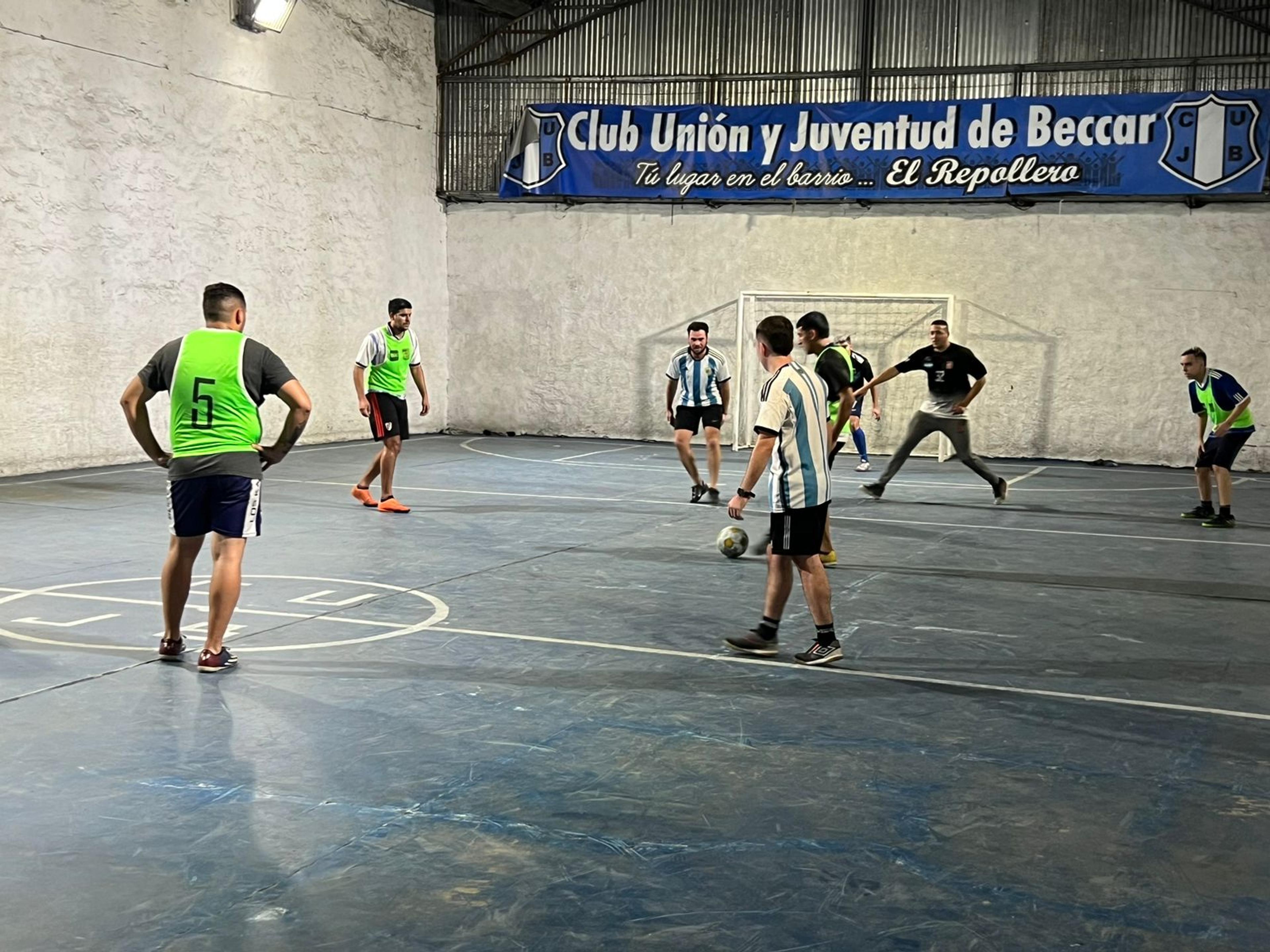 People in Argentinian football tops and training bibs play soccer in an indoor arena. It is the Argentinian street soccer team.