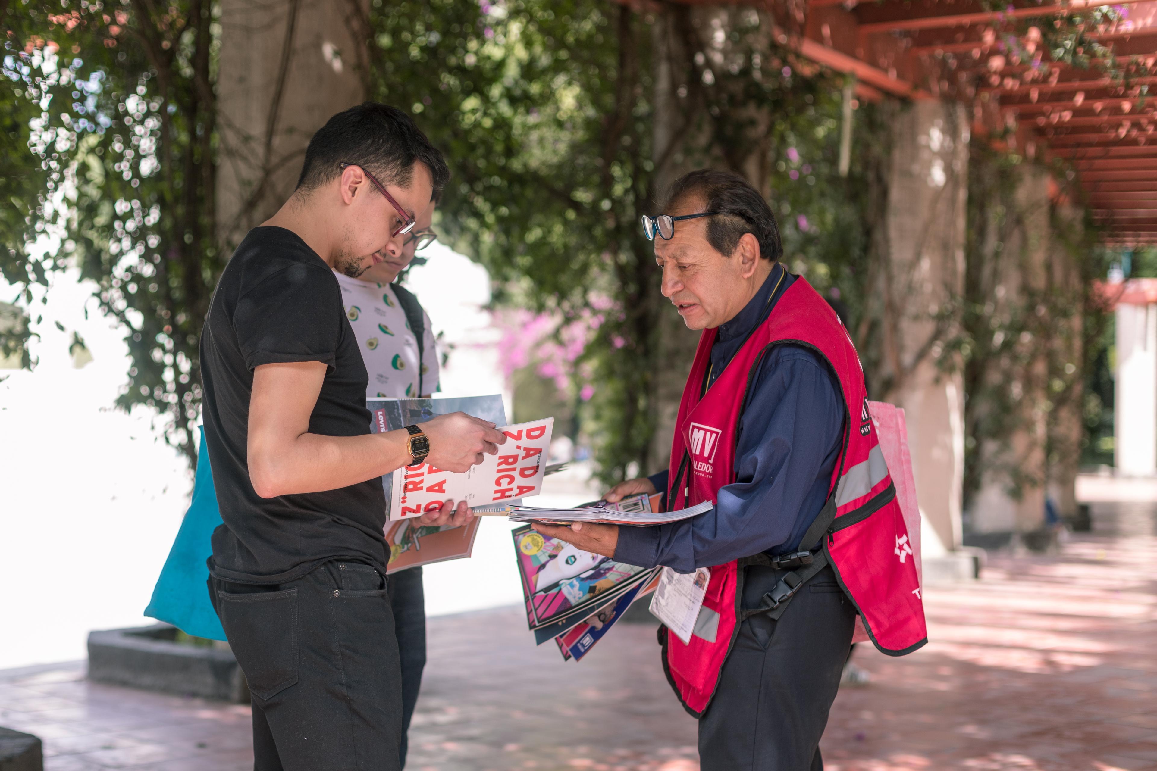 A man in a red vest sells a street paper to a passerby