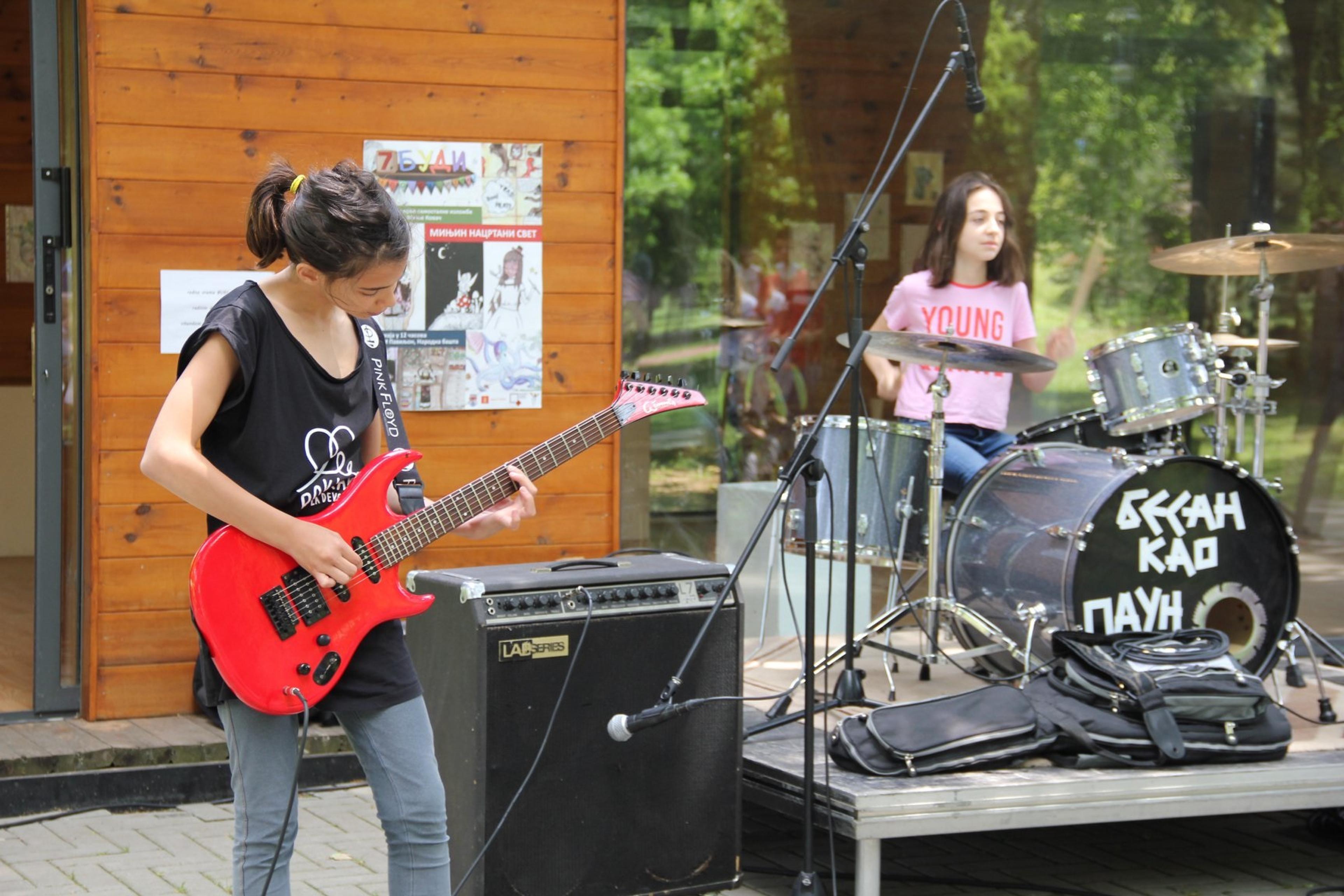 two young girls in front of a log cabin, one is playing the drums, the other a red guitar