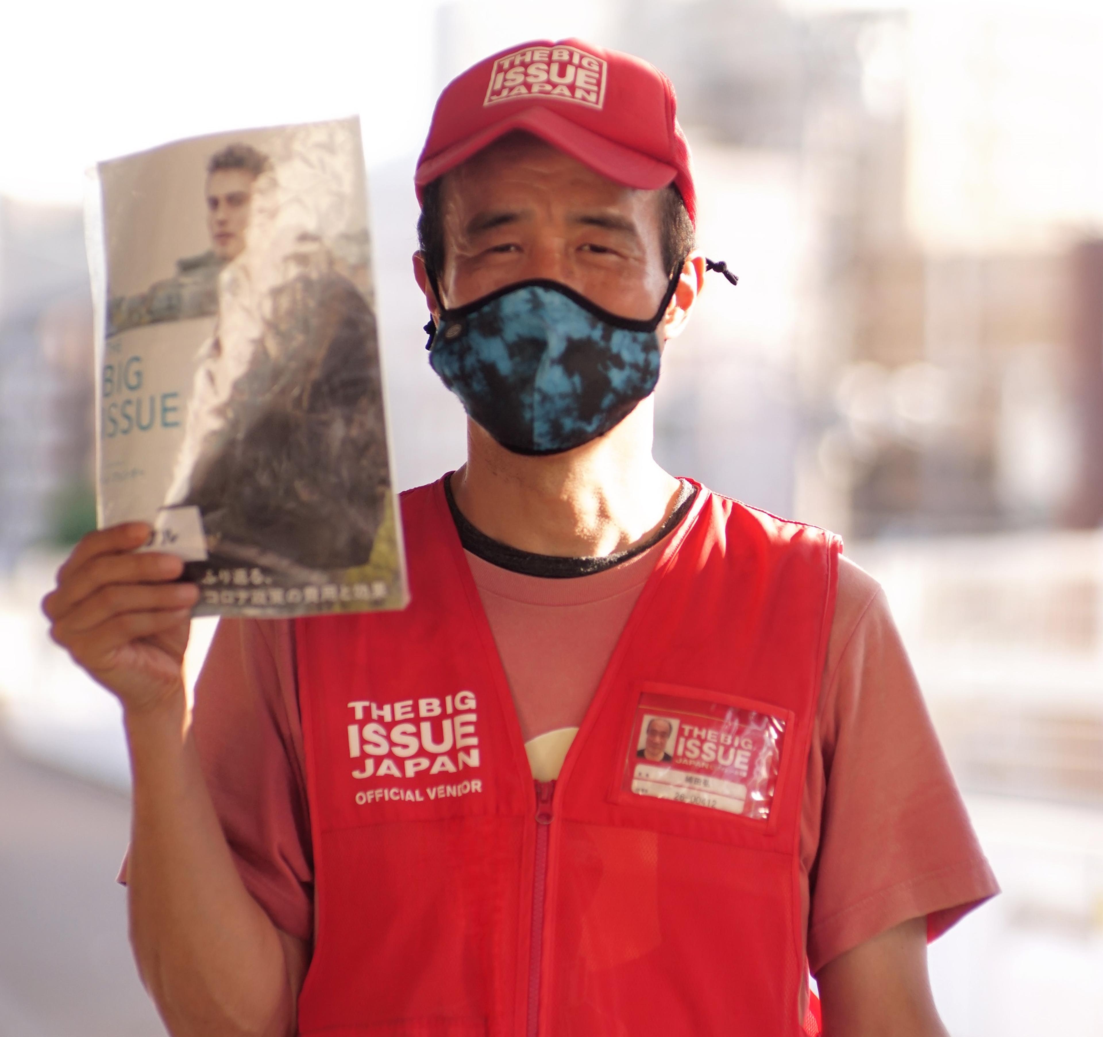 Man with a red hat and vest and wearing a mask holds a copy of The Big Issue Japan magazine