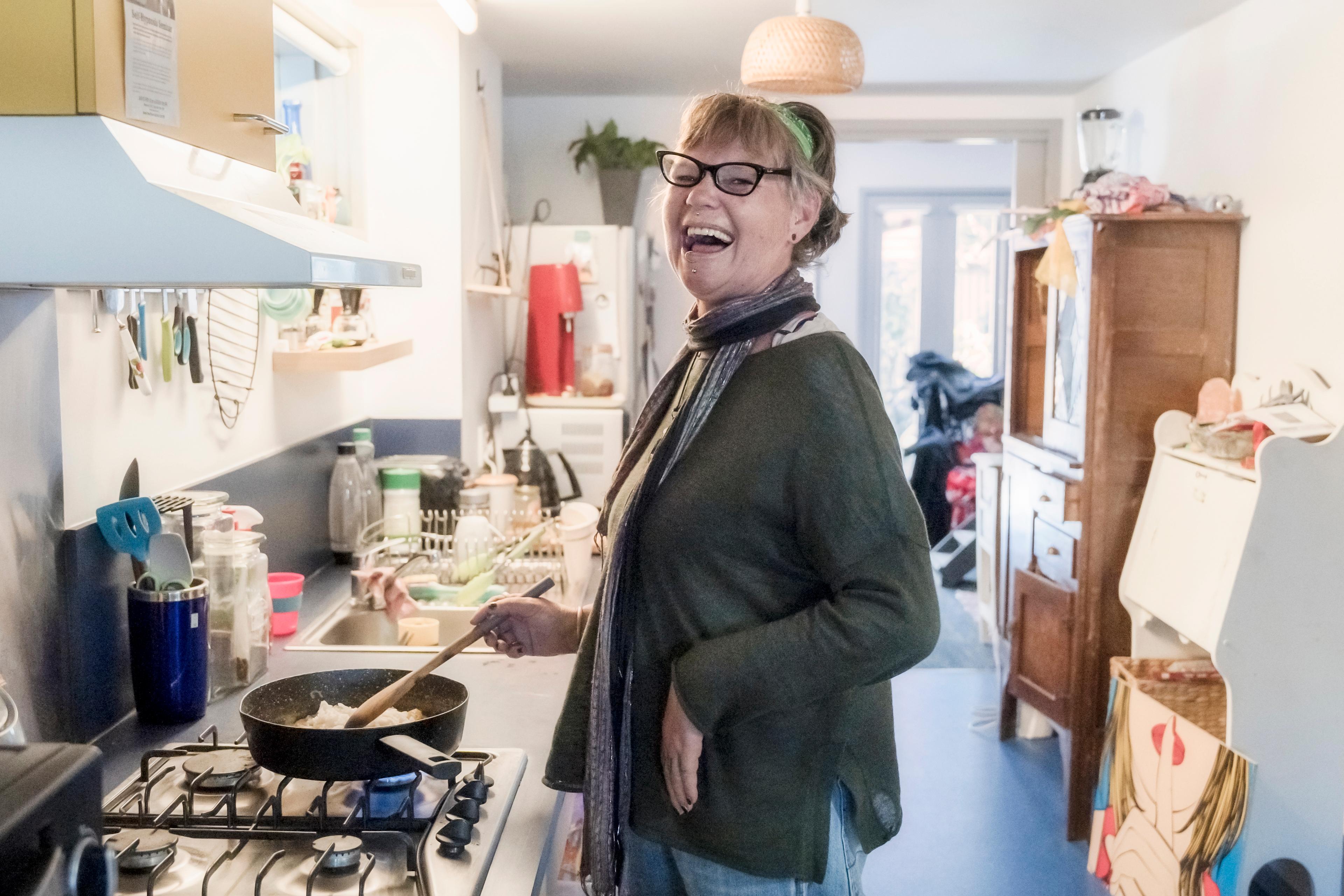 Danni stands in a kitchen laughing. She is wearing a scarf and glasses.