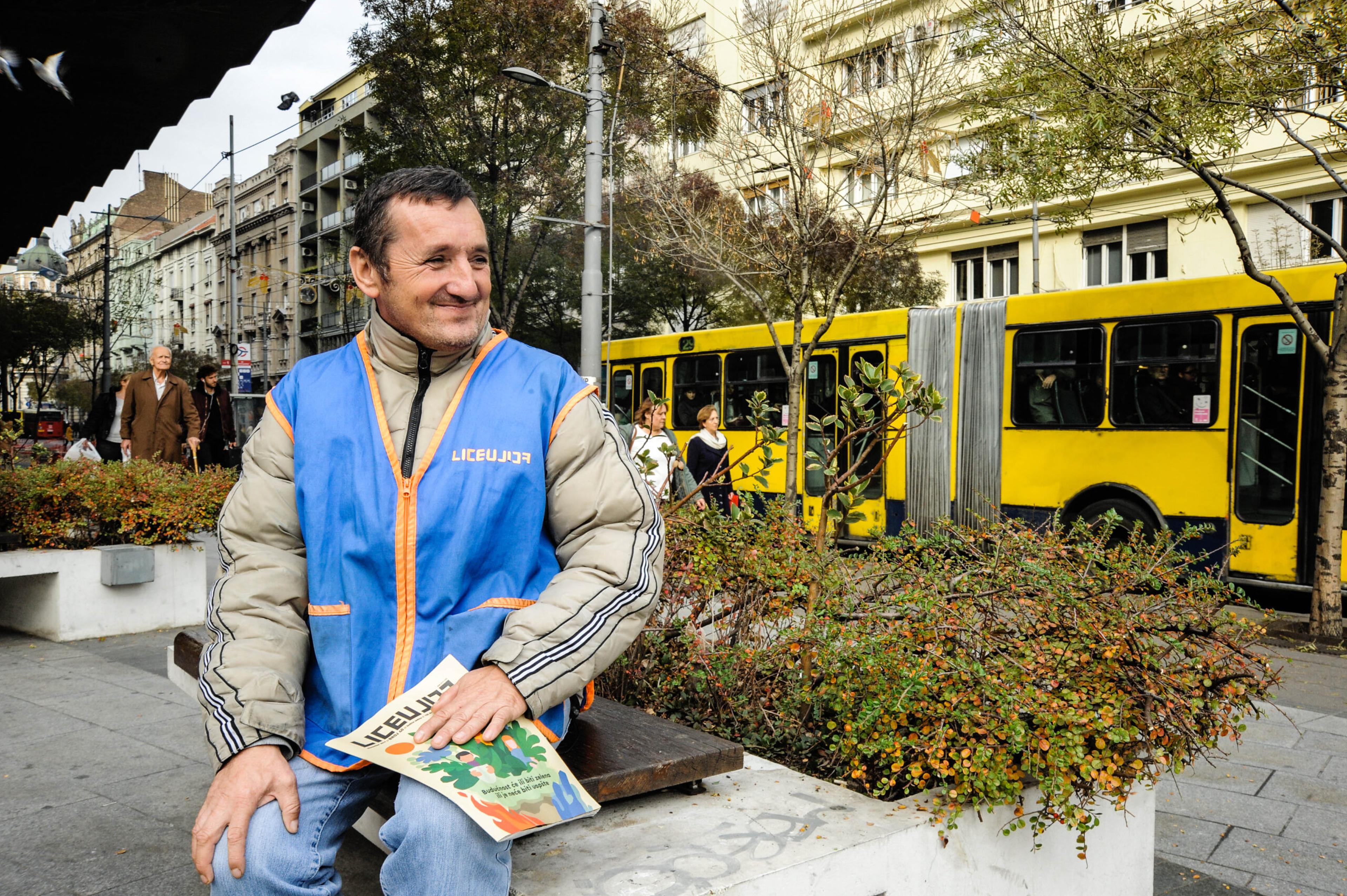 Liceulice Tomislav sits on a bench smiling, wearing his blue and orange Liceulice vest and holding the magazine. He is surrounded by trees and plants, and there is a yellow bus behind him.