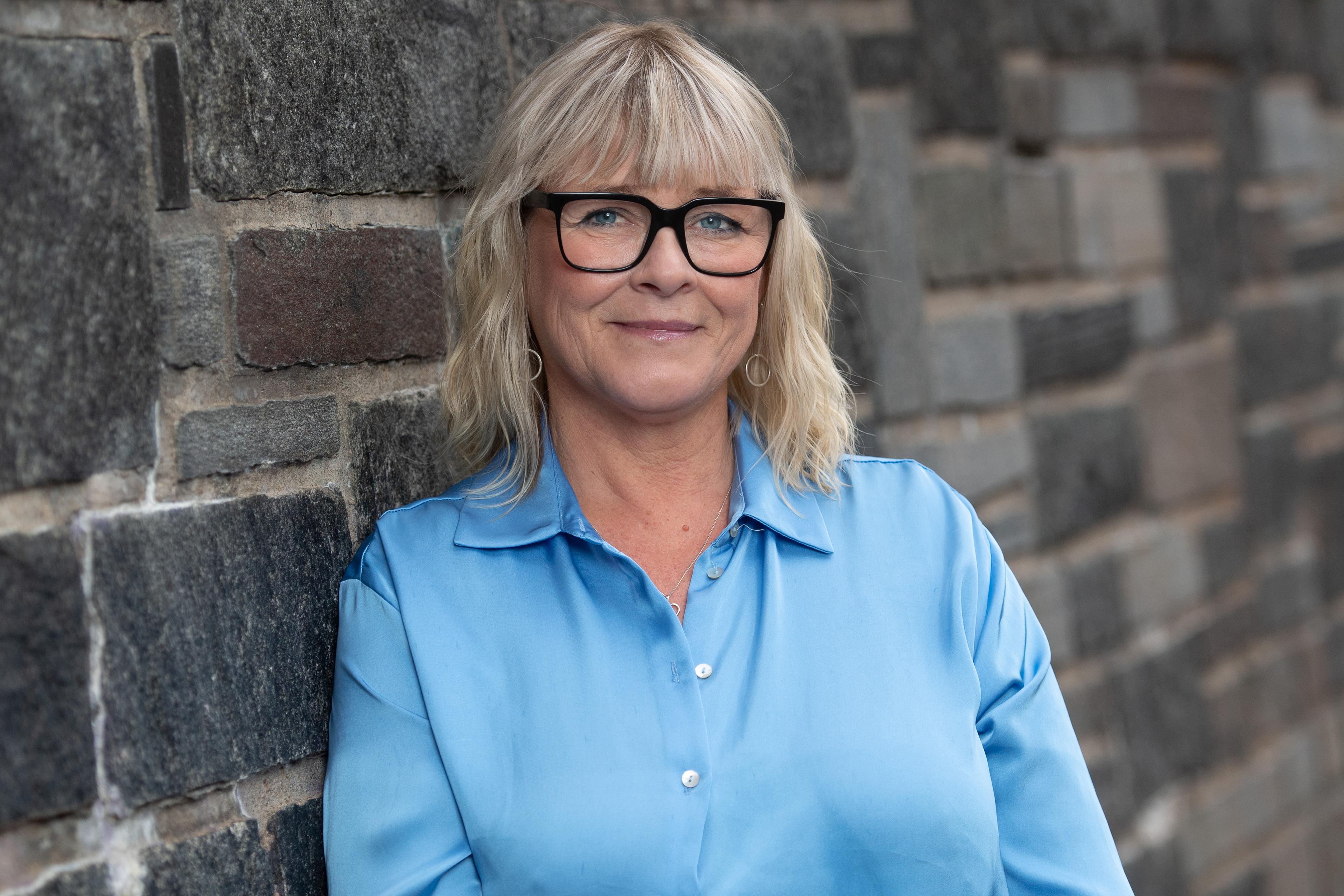 Sarah Britz stands against a brick wall with blonde hair, thick black glasses and a blue shirt.