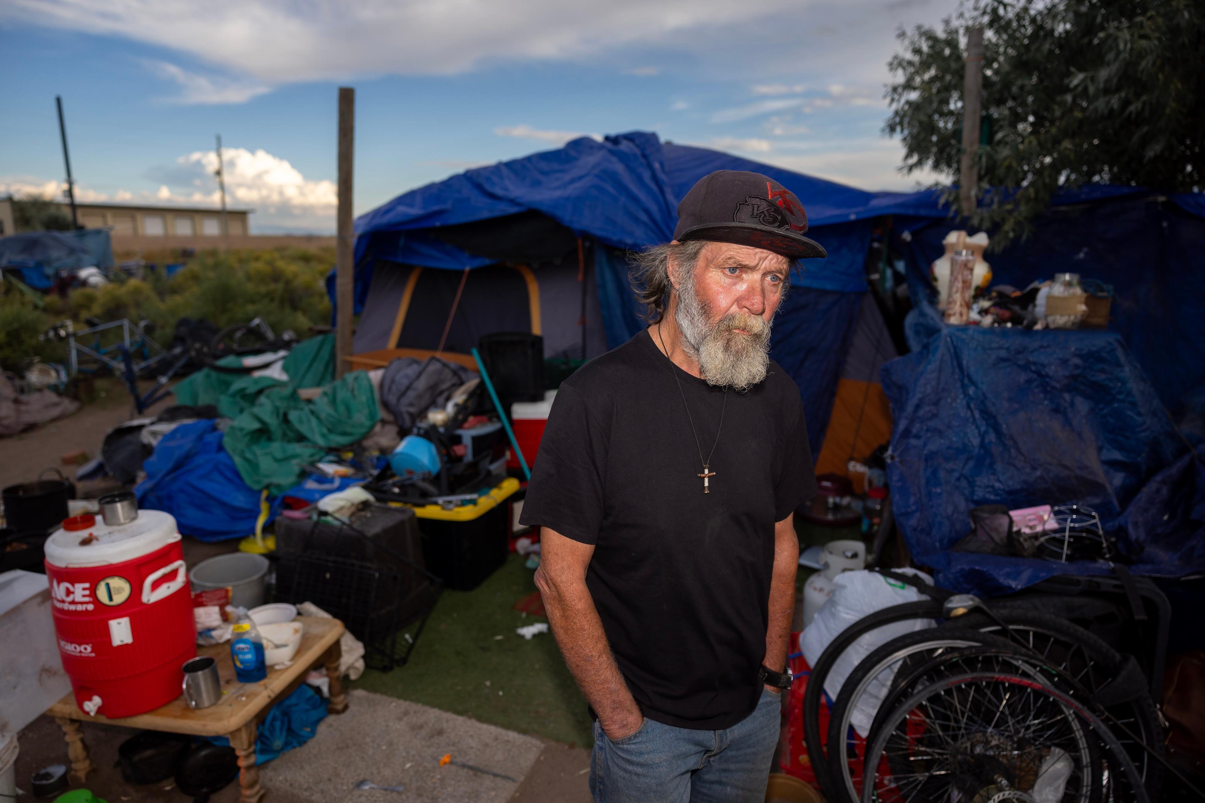 a bearded man standing in front of a blue tent and a pile of supplies