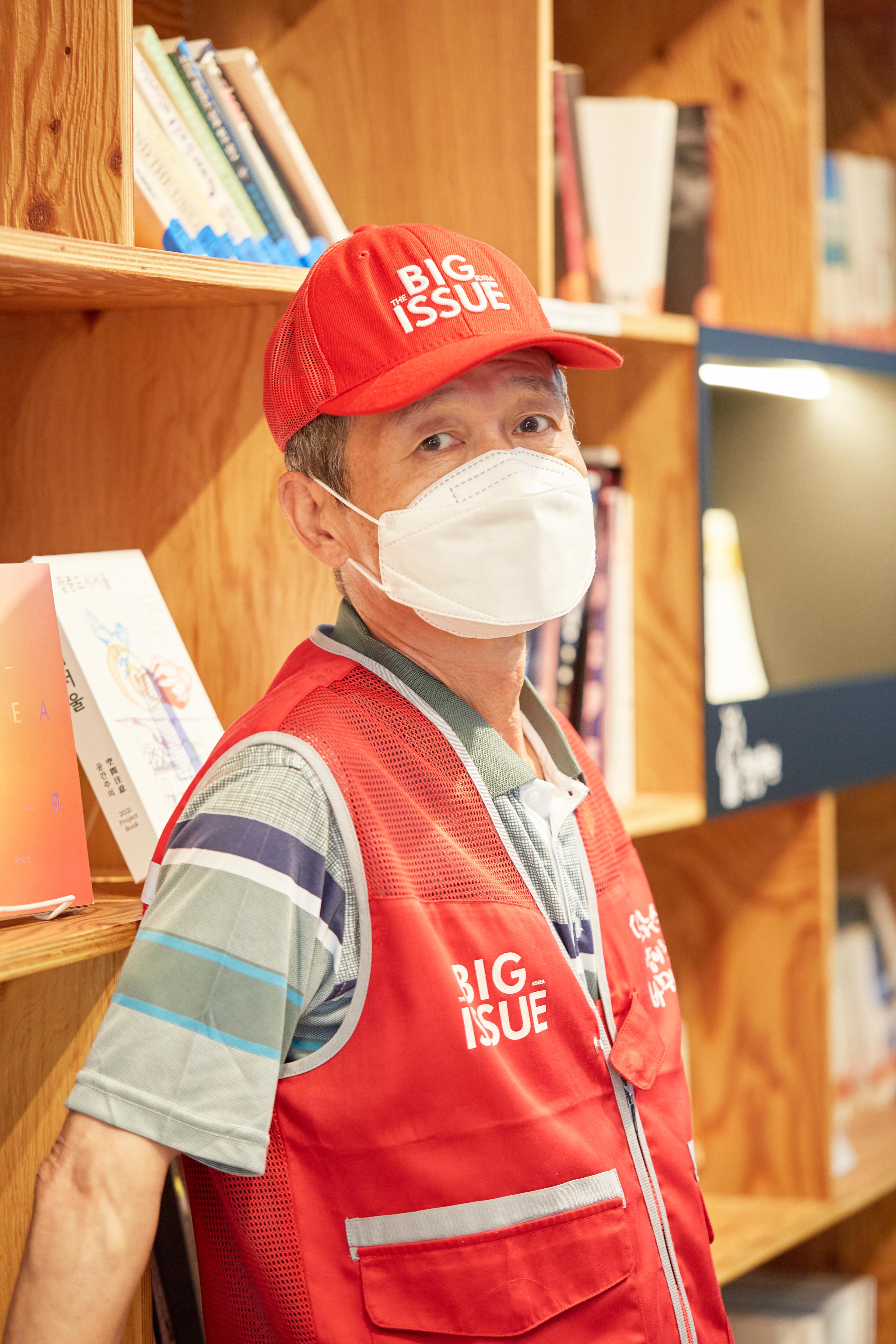 A man in a red hat and mask leans against a bookcase