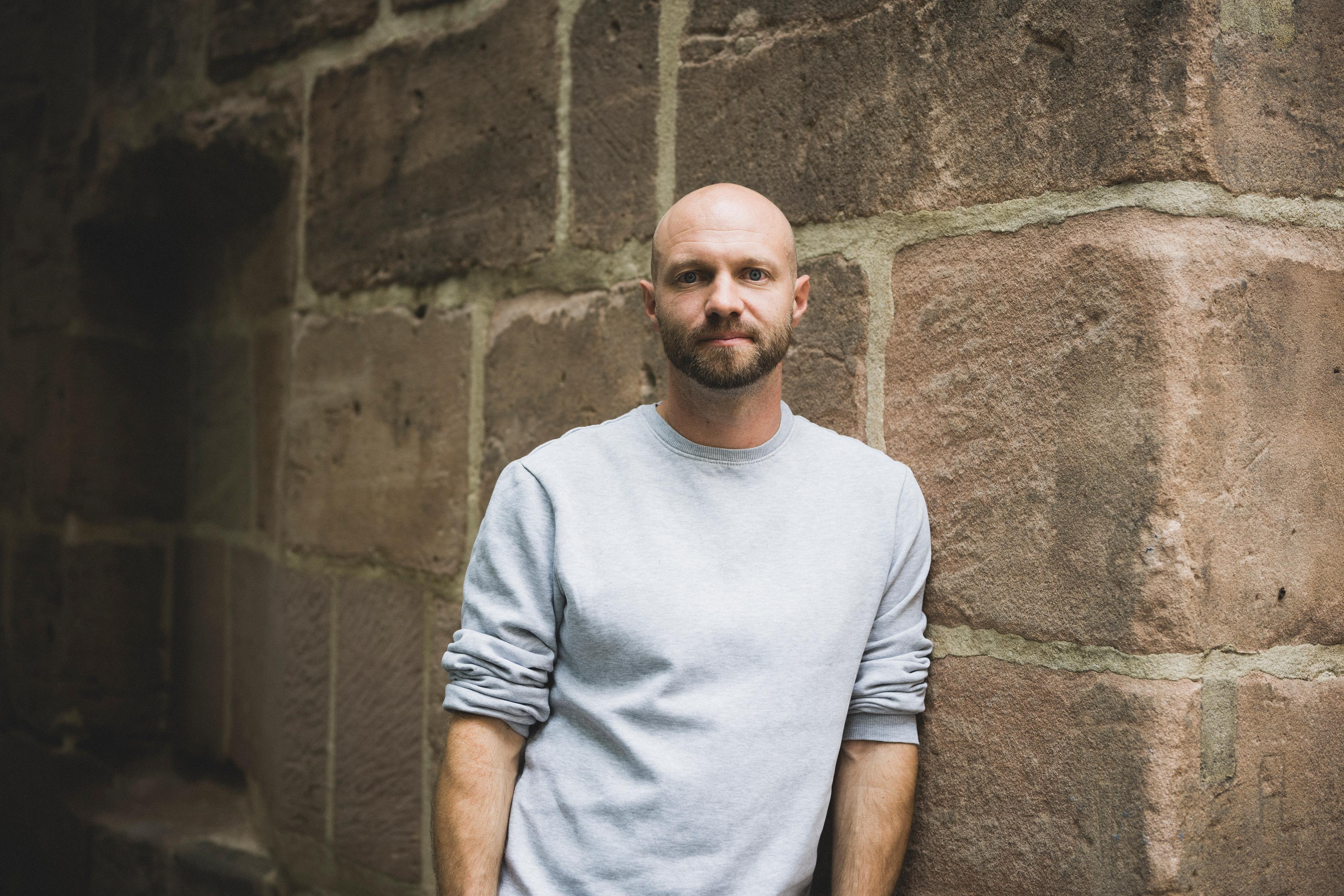 Political scientist Martin Stammler leans against a wall. He is bald with a beard and is wearing a grey sweater.