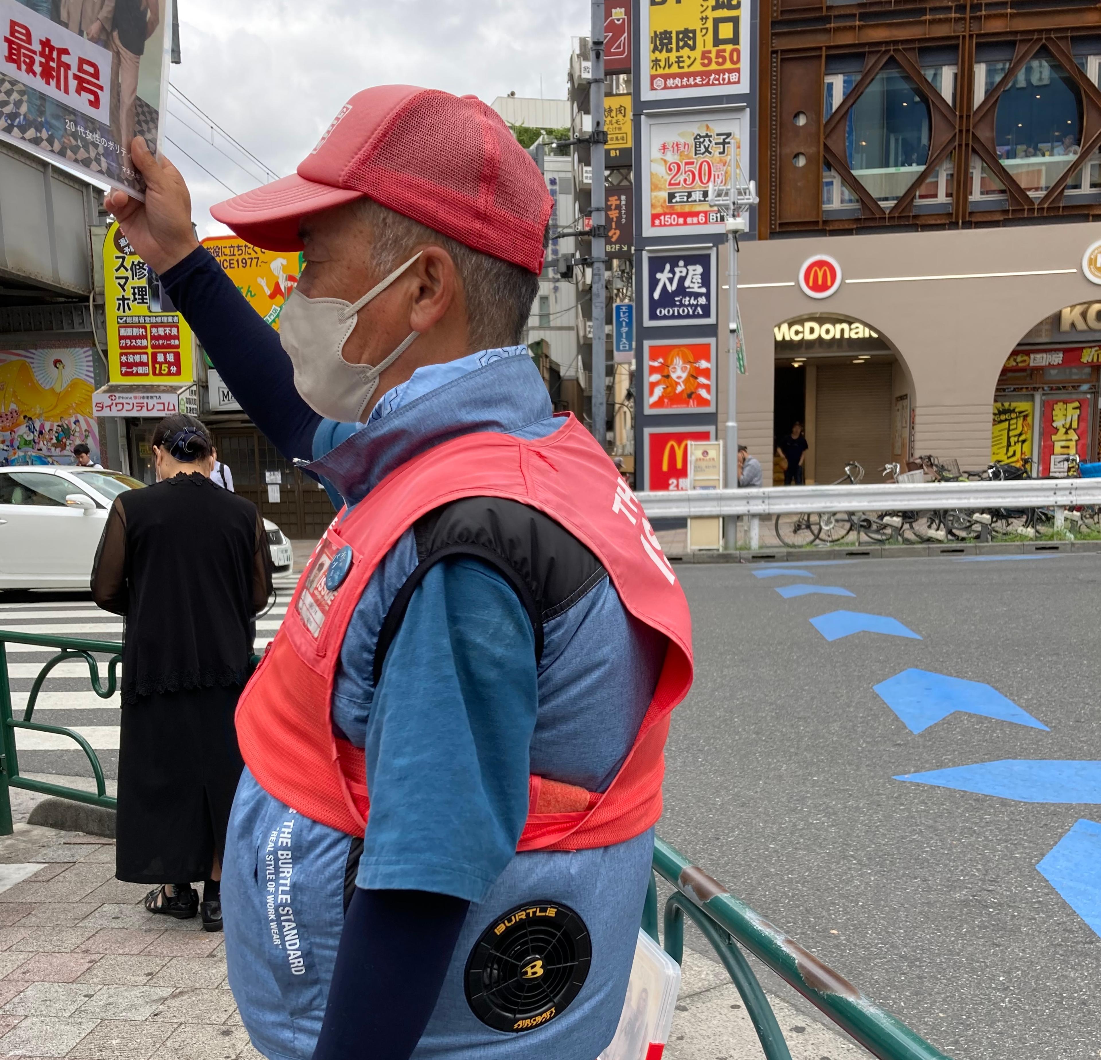 A street paper vendor of The Big Issue Japan stands on the street in the heat wearing a jacket that regulates air to cool the wearer down.