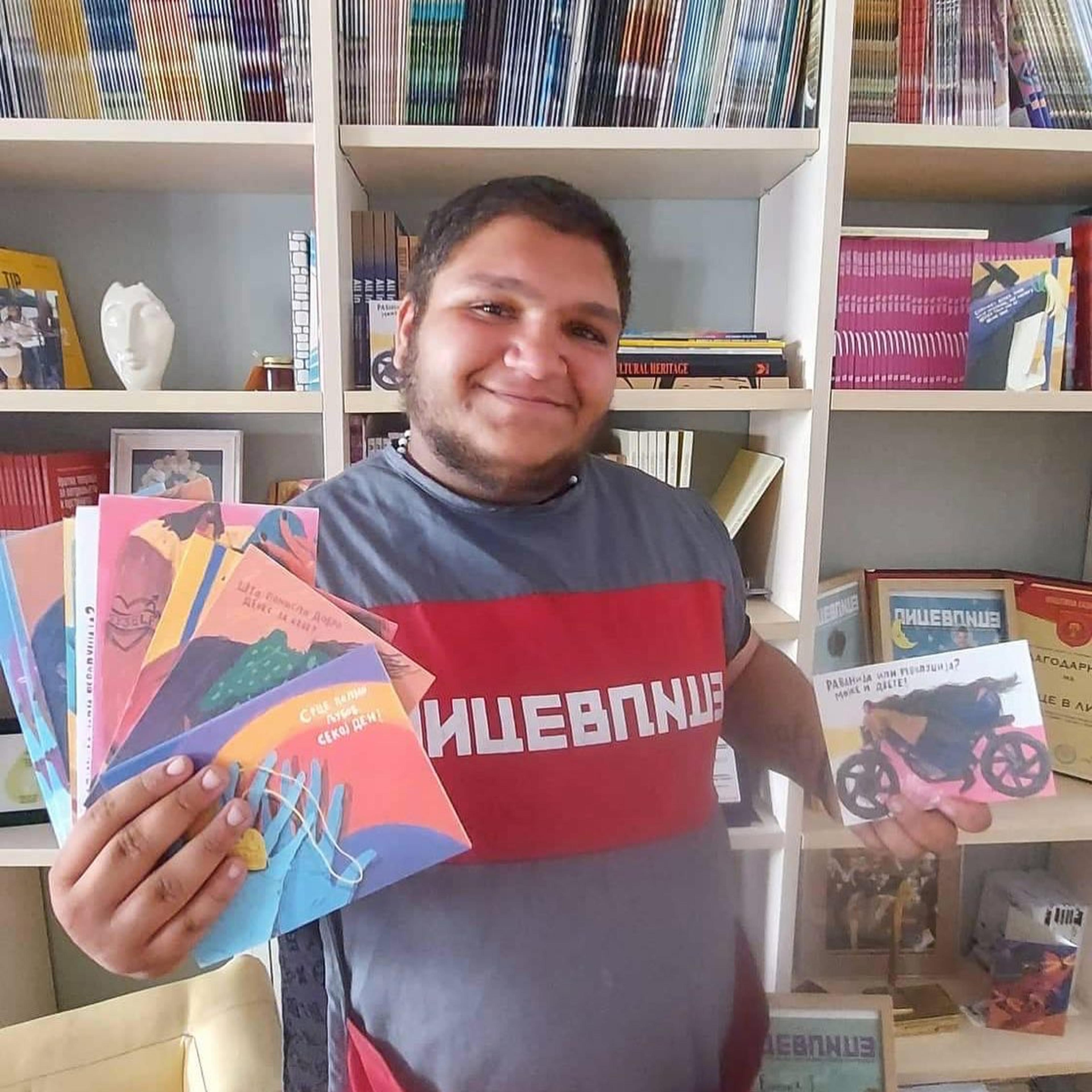 Memet holds books in his hands, smiling, wearing a red and grey Lice v Lice t-shirt.