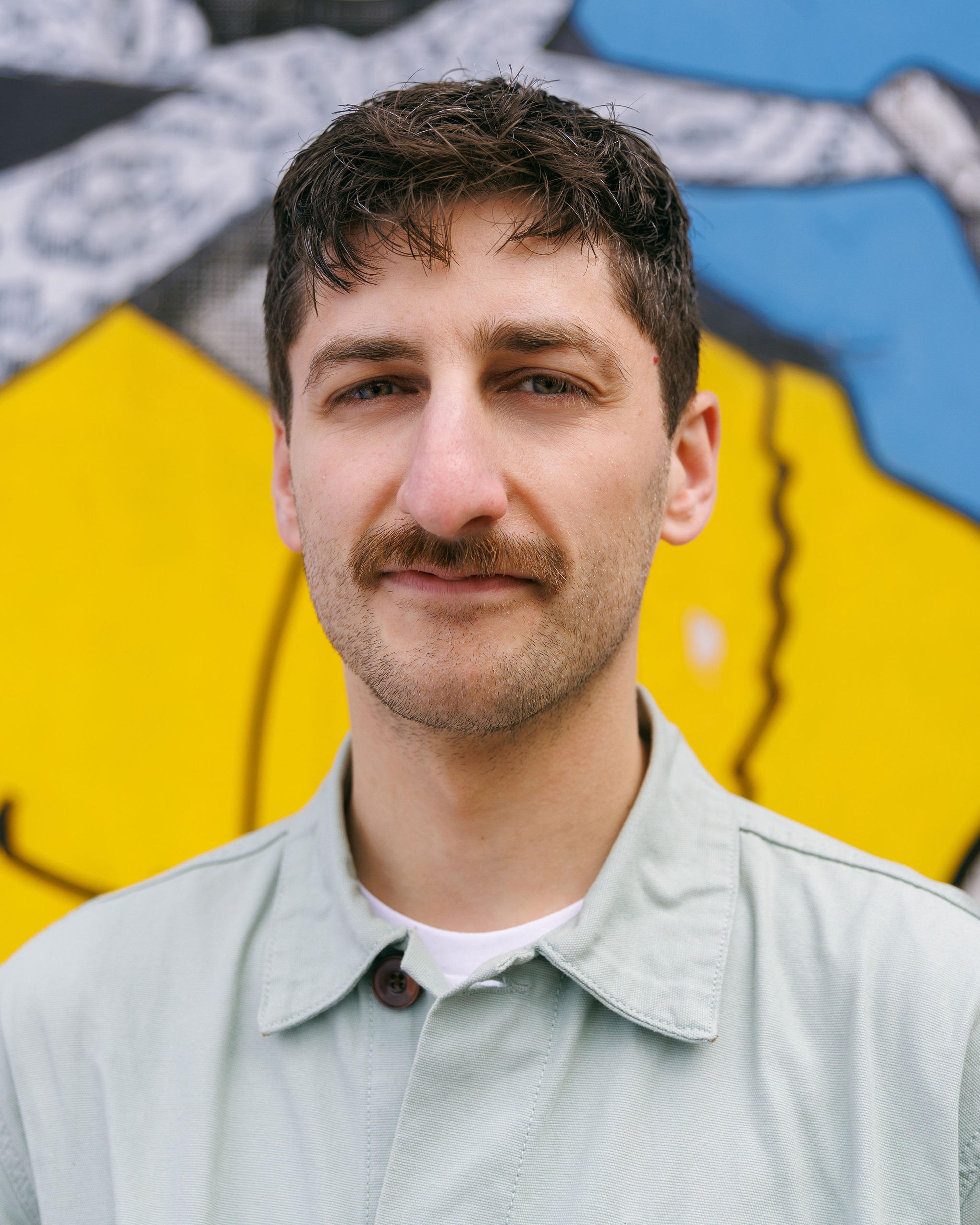 A headshot of INSP editor Tony Inglis, wearing a teale jacket in front of yellow and blue street art