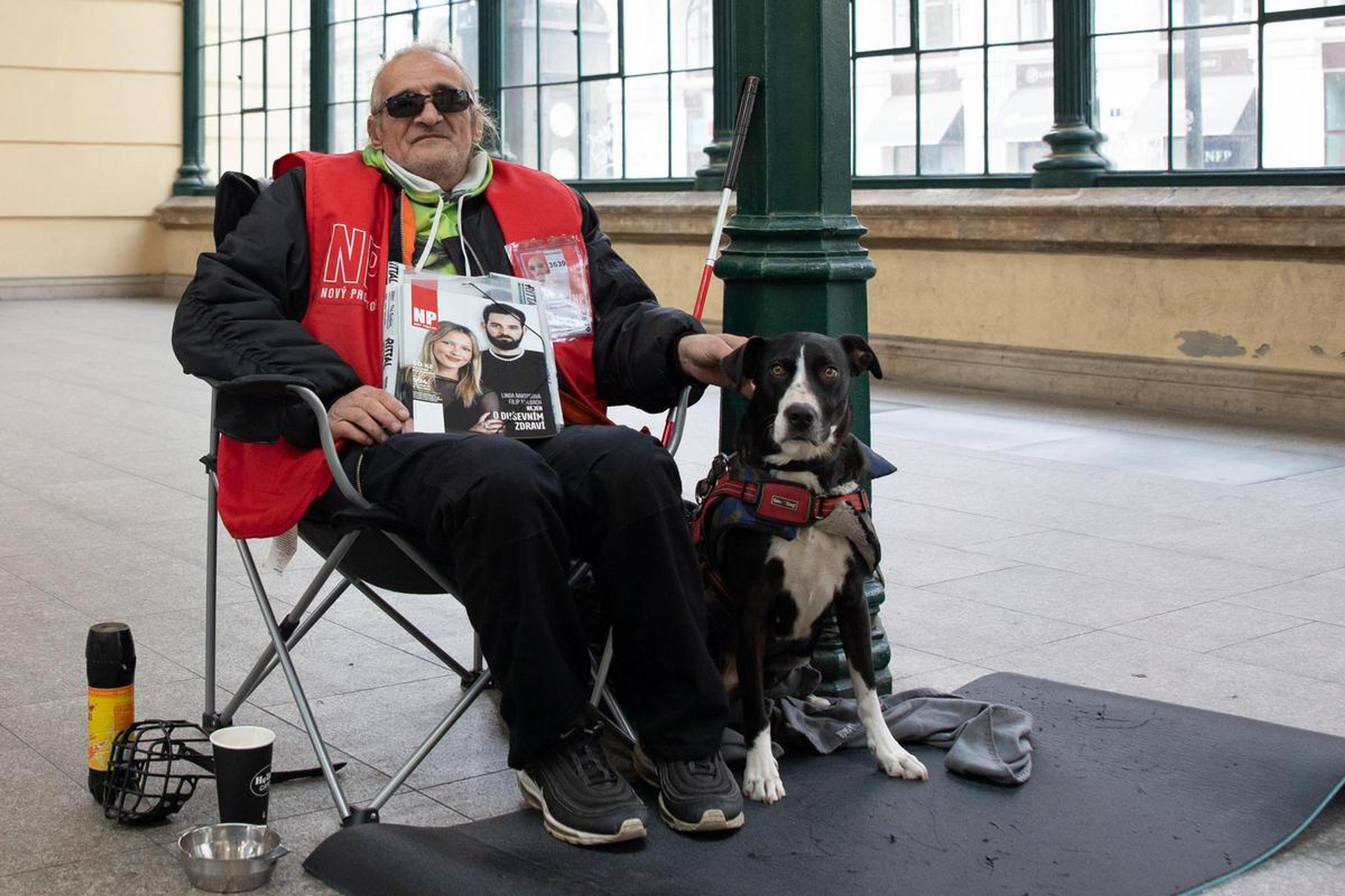 Vendor Josef sits in a chair next to his dog Kyra. Kyra is black and white, and Josef is wearing a red vest and sunglasses.