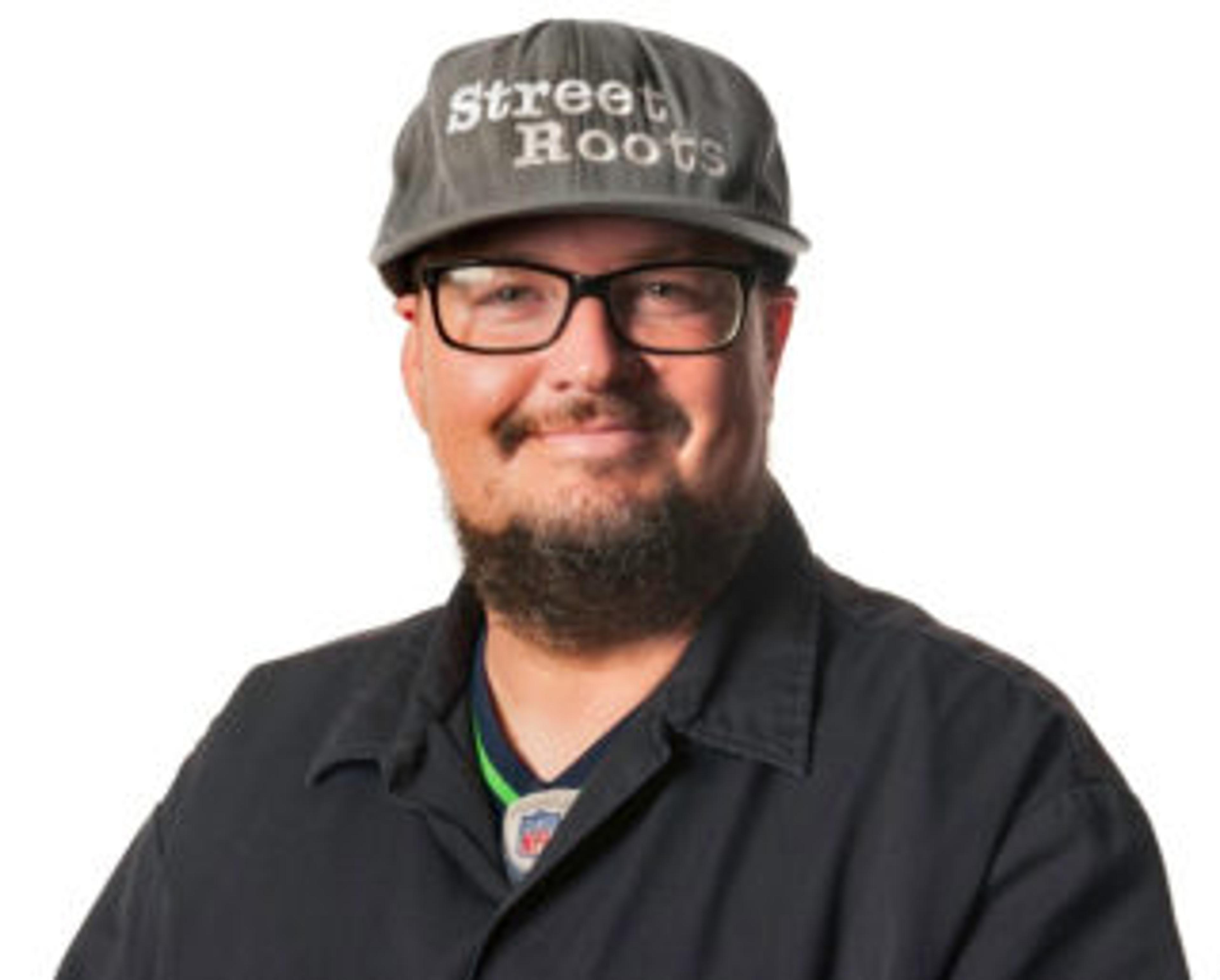 A headshot of INSP North America director Israel Bayer, wearing a black shirt, glasses and a cap with the logo of Portland street paper Street Roots