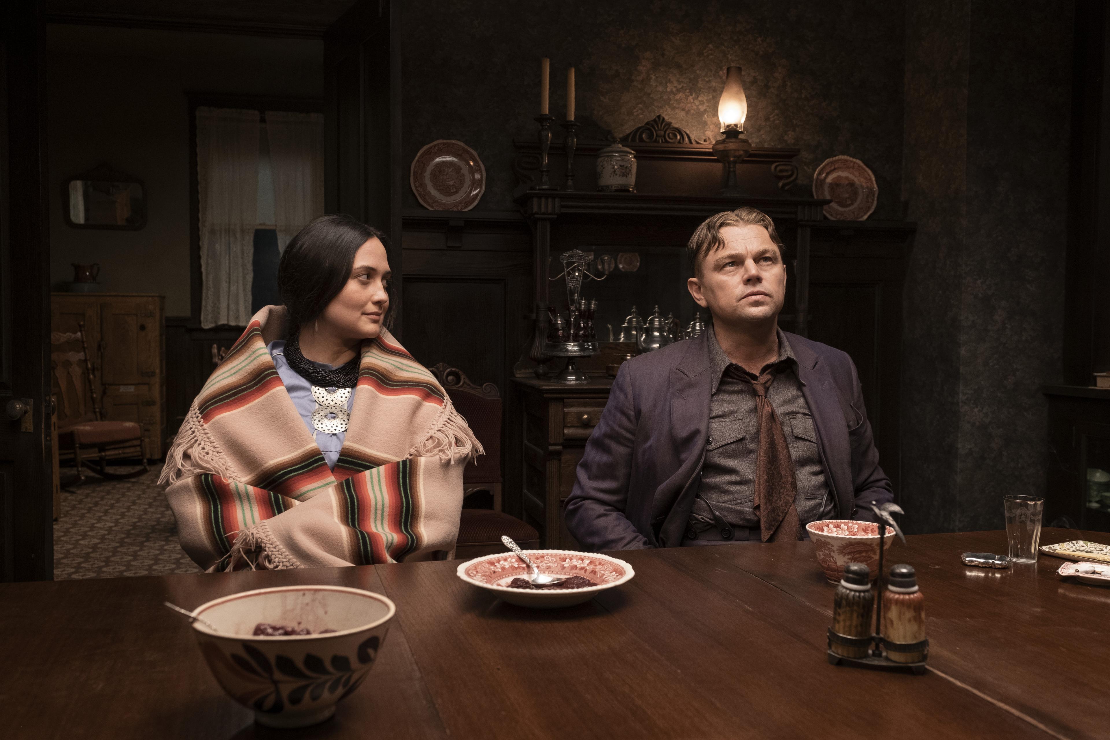 Lily Gladstone and Leonardo DiCaprio act on the set of the film Killers of the Flower Moon. They are in period costume and are sat at a dining table.