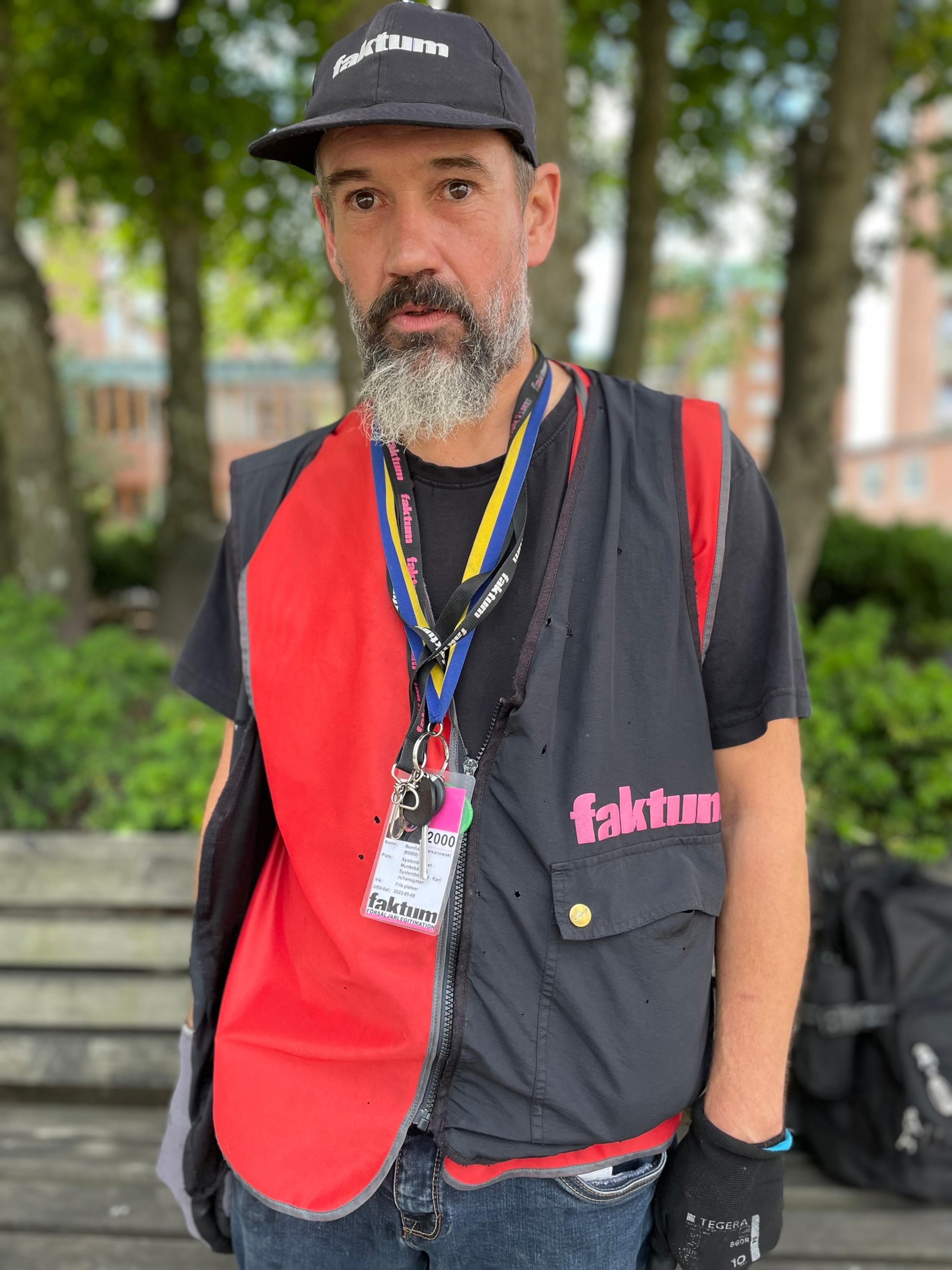 A vendor of the Swedish street paper Faktum with a grey beard wearing a Faktum branded vest and hat.