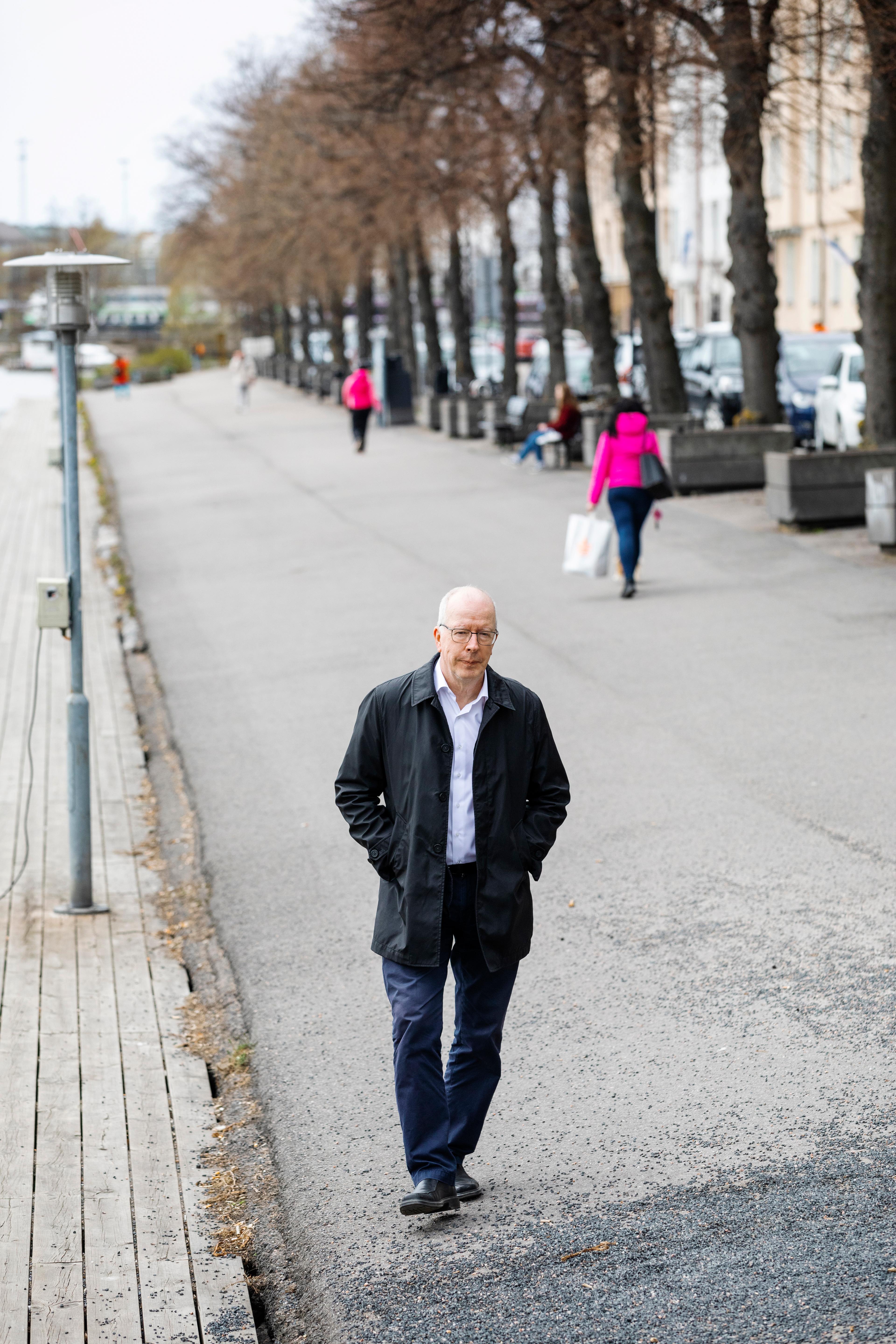 Juha Kaakinen wears glasses a white shirt and black jacket. He is photographed walking down a busy street.