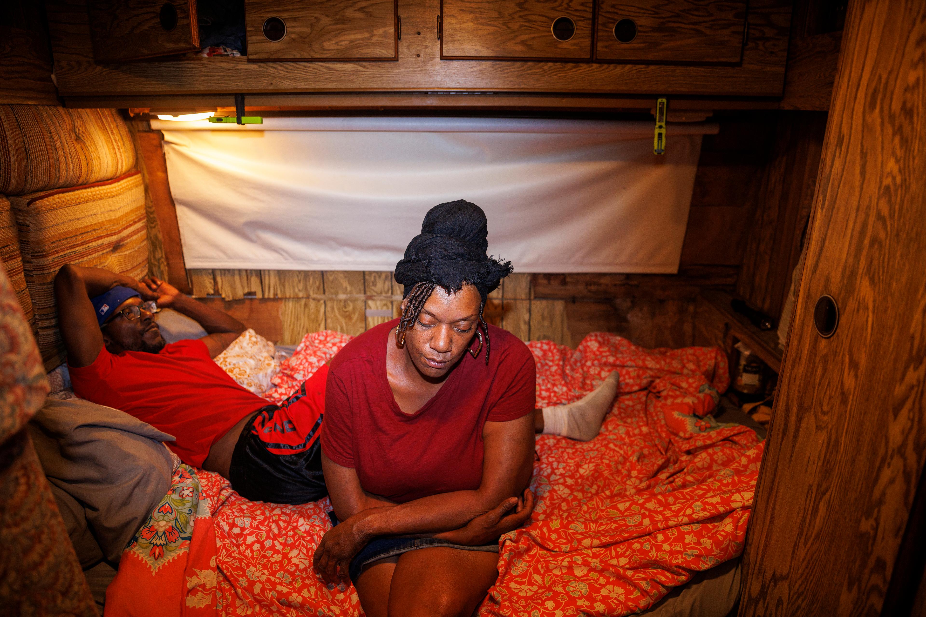 A woman sits on a bed in front of a man lying down, in a camper van.