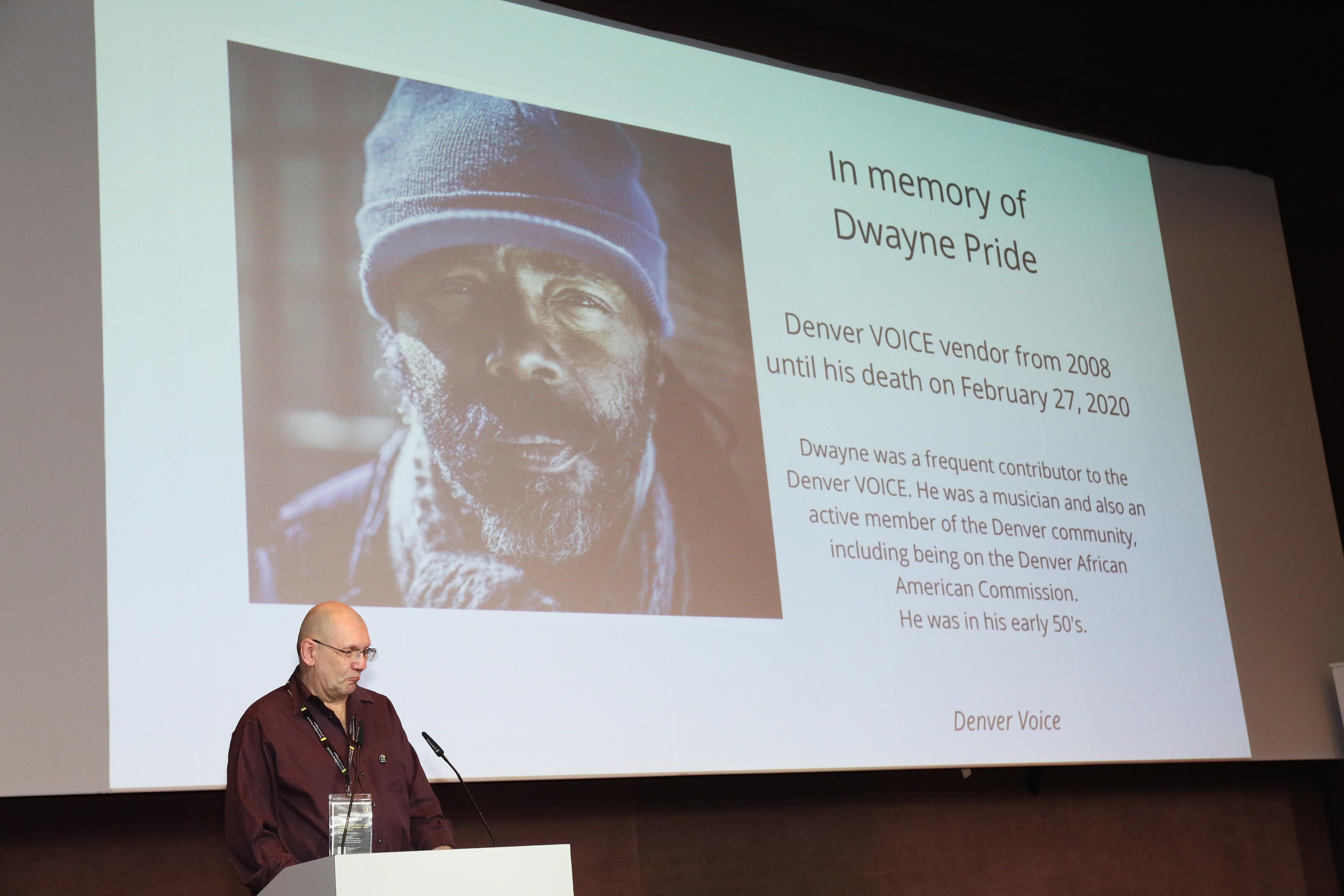 A man presents behind a lectern and in front of a screen where street paper vendors who have died are memorialised.