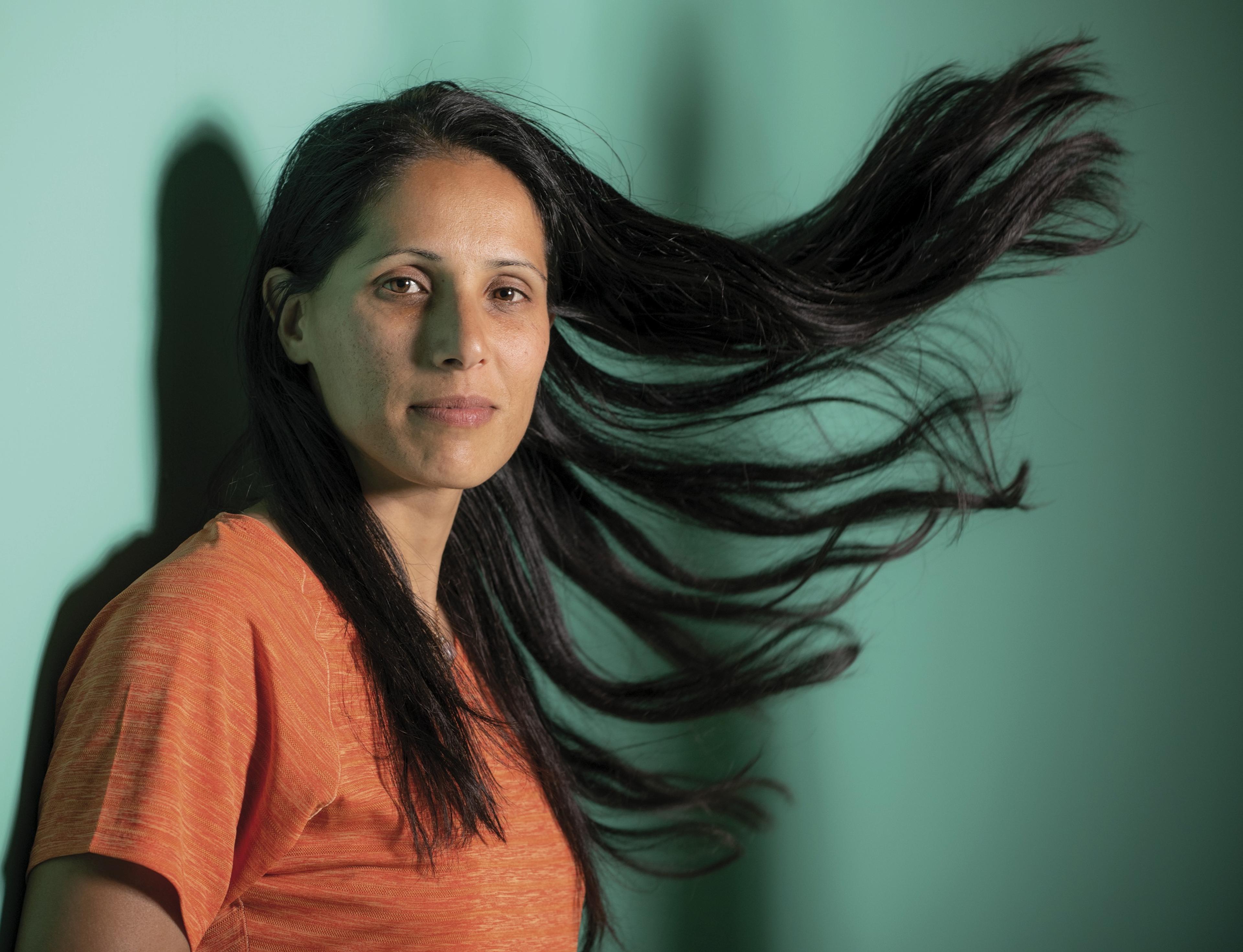 a woman in an orange top leans against a green wall with her hair blowing in the wind