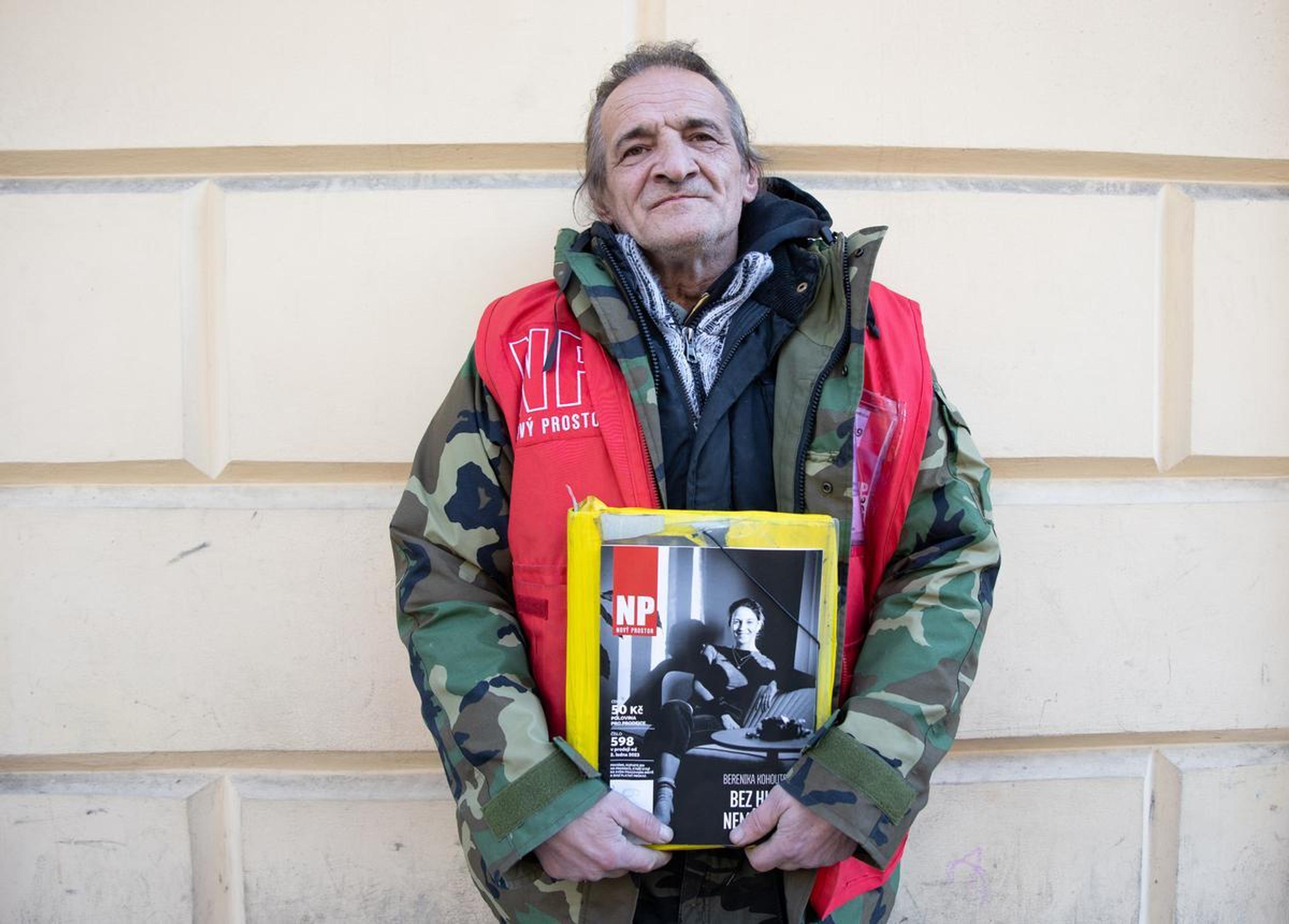 Josef stands against a wall wearing a cargo jacket and red vest holding a copy of the Novy Prostor Czech street paper