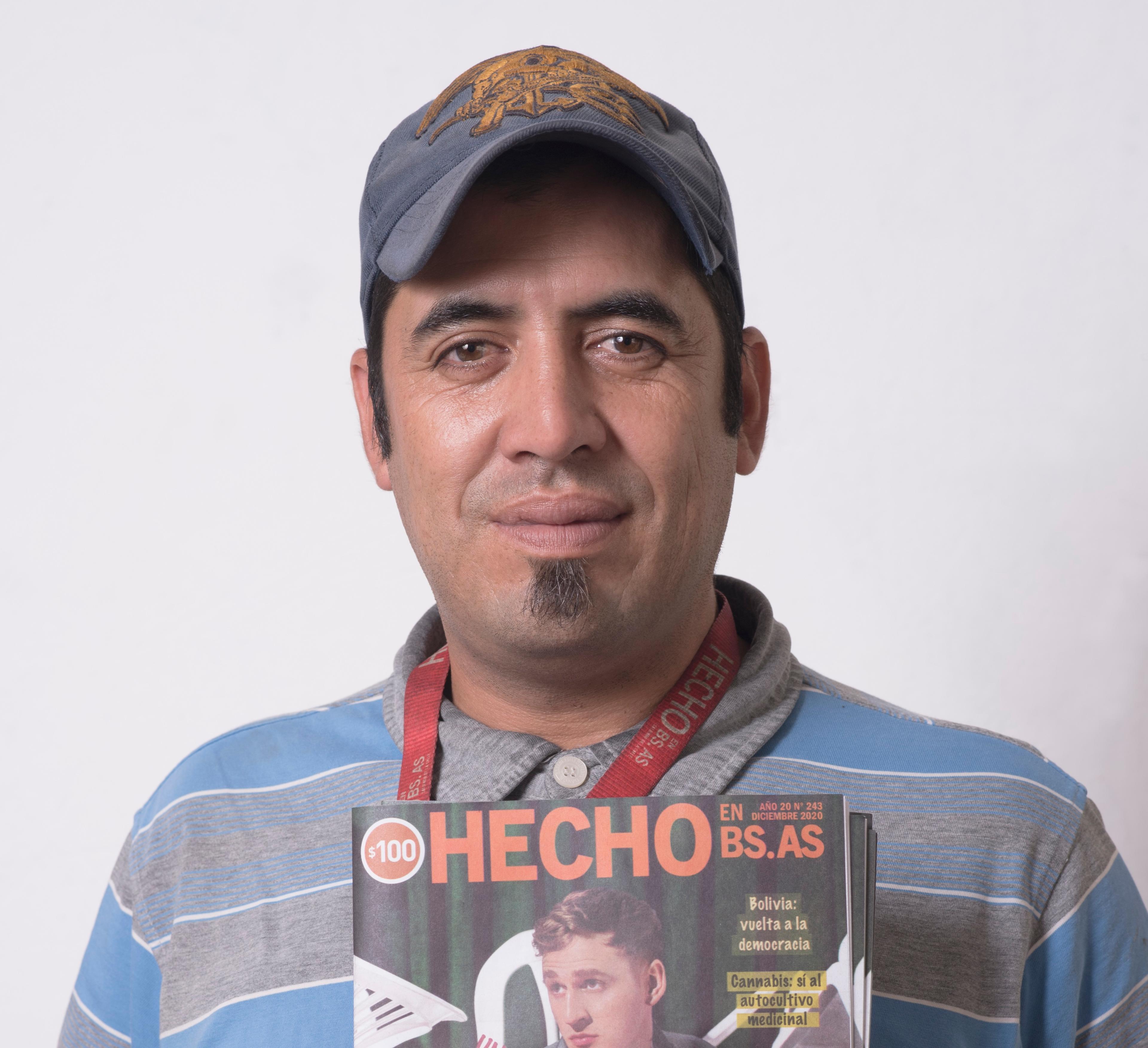 Hecho vendor Luciano is wearing a blue cap and stripy polo top and is holding a copy of the Hecho en Bs. As. street paper