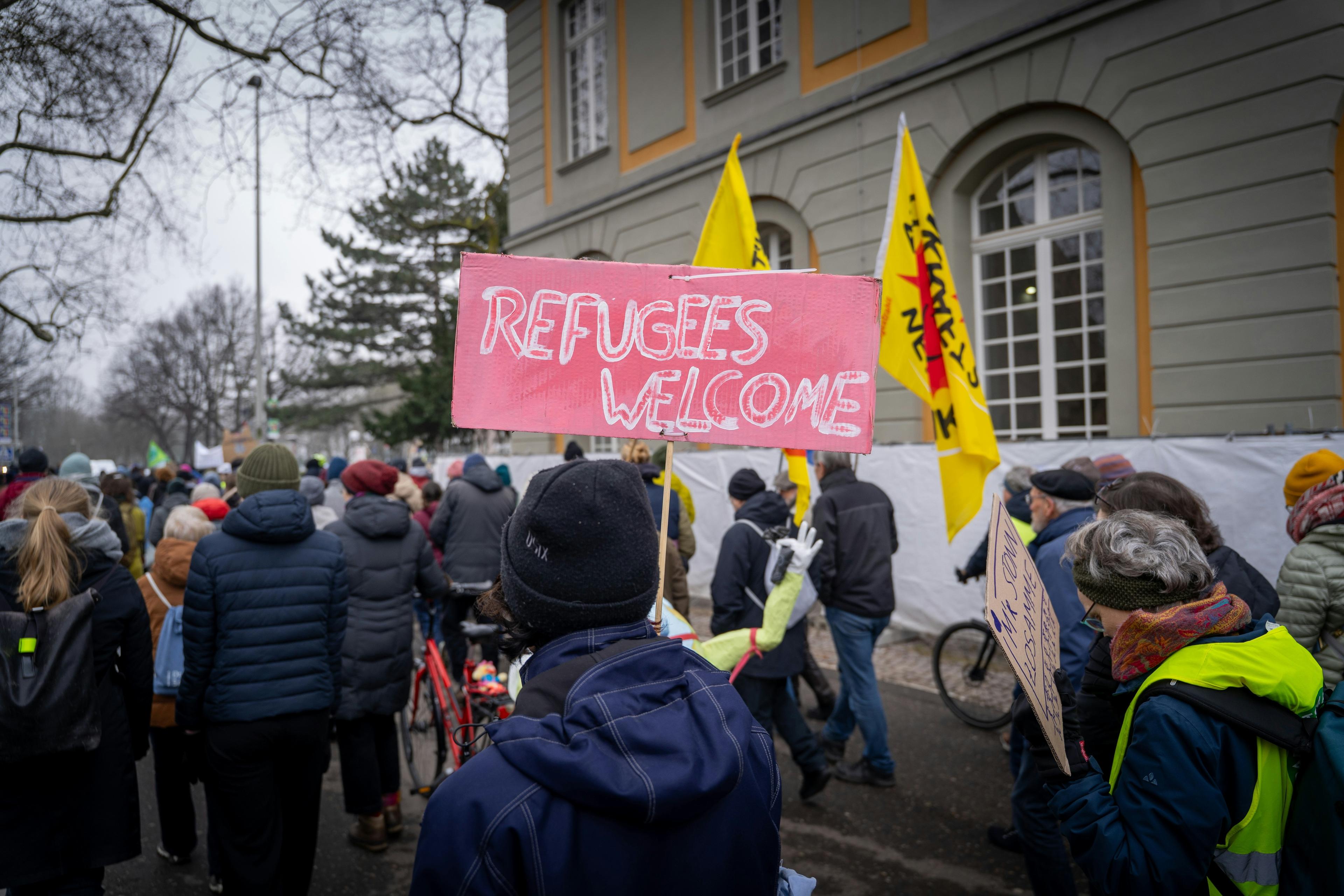 a man taking part in a march, holding a pink placard that says 'refugees welcome'