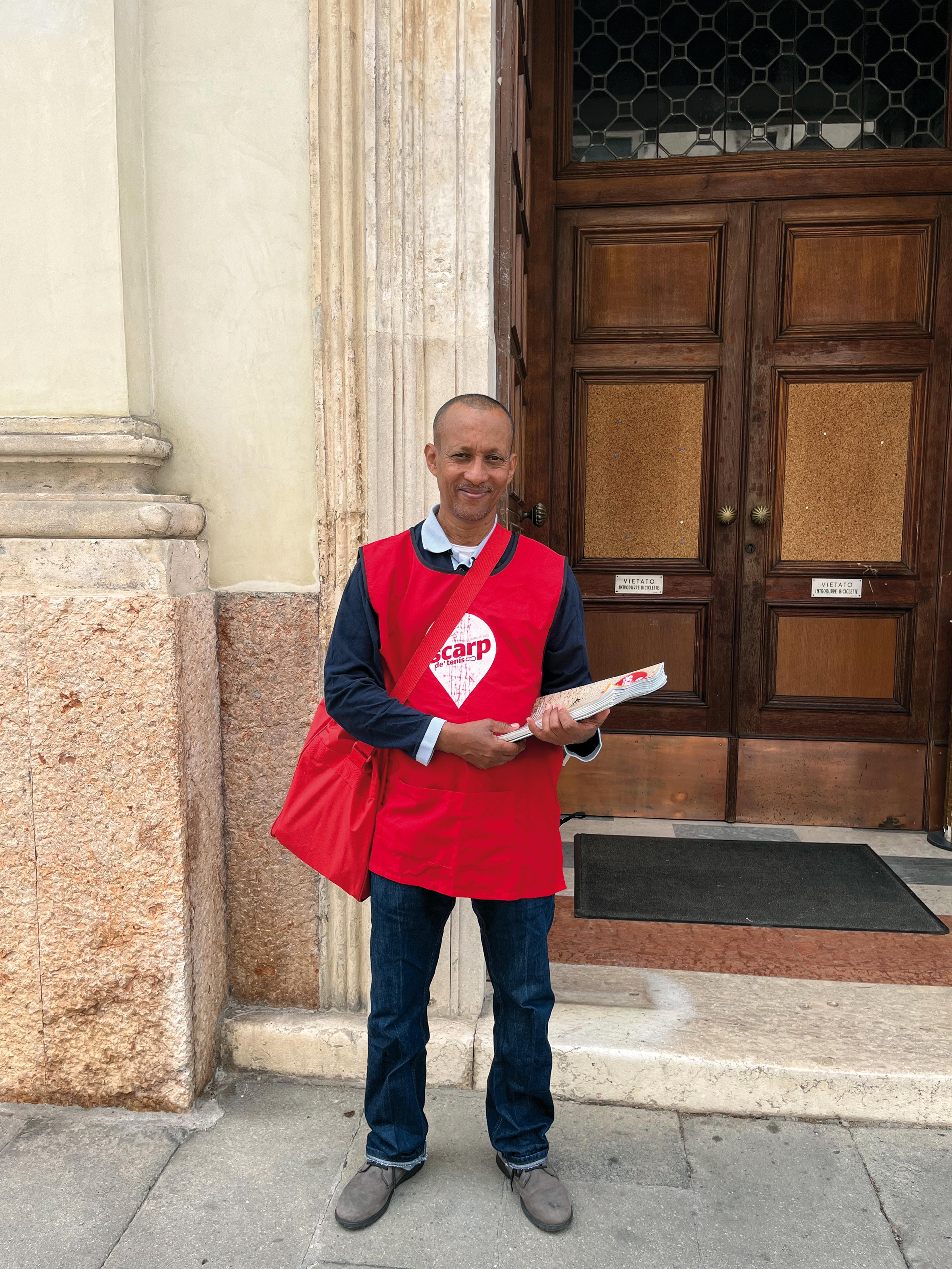 Scarp de' tenis vendor Giorgio stands in front of a door wearing a red vest and holding a red bag with the Scarp logo. He is holding a bundle of magazine.