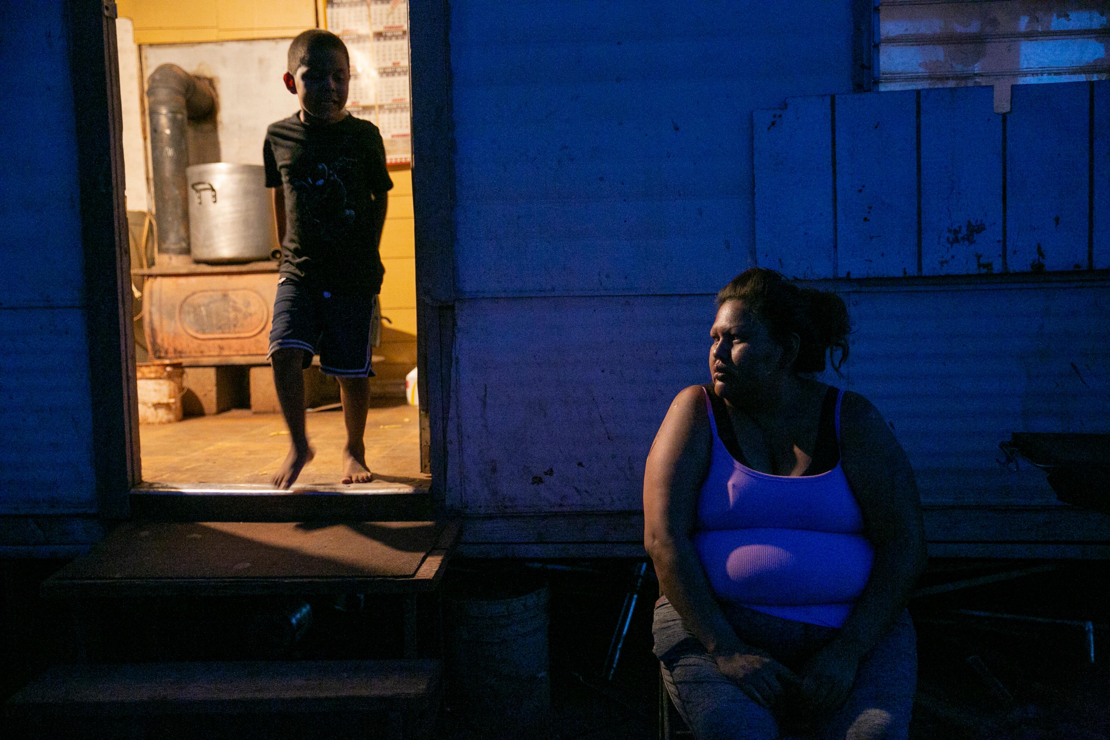 A woman sits outside her house in the darkness, while her sun stands in the doorway lit from behind.