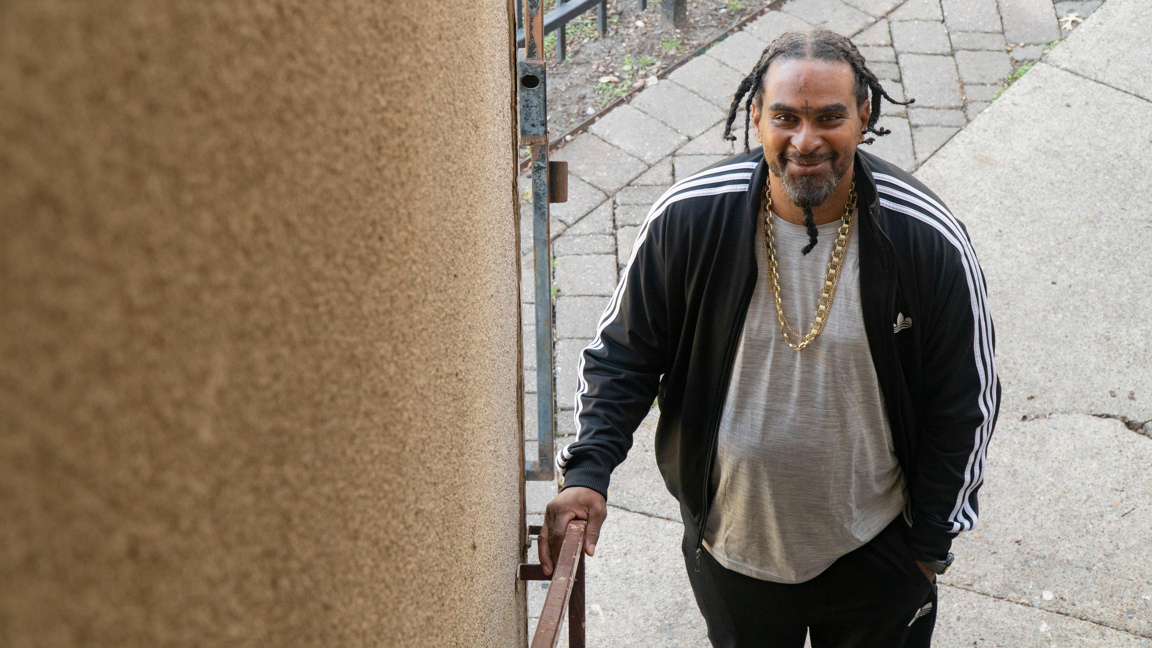 Darin Thomas stands at the foot of a staircase. His hair is braided and is wearing a black jacket, a grey t-shirt and a gold chain.