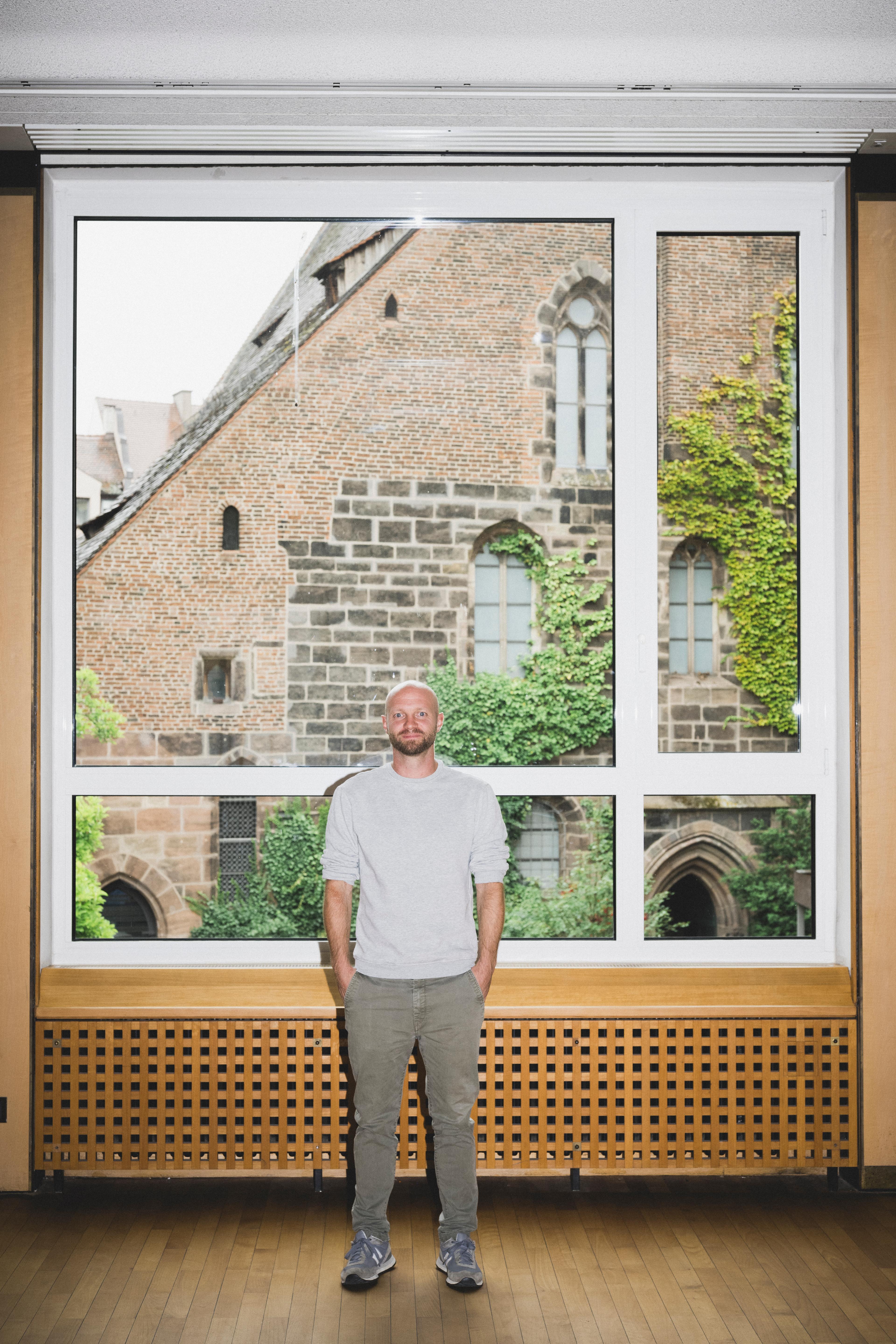 Political scientist Martin Stammler stands in front of a window with his hands in his pockets. He is bald with a beard, and is wearing a grey sweater and khaki trousers.