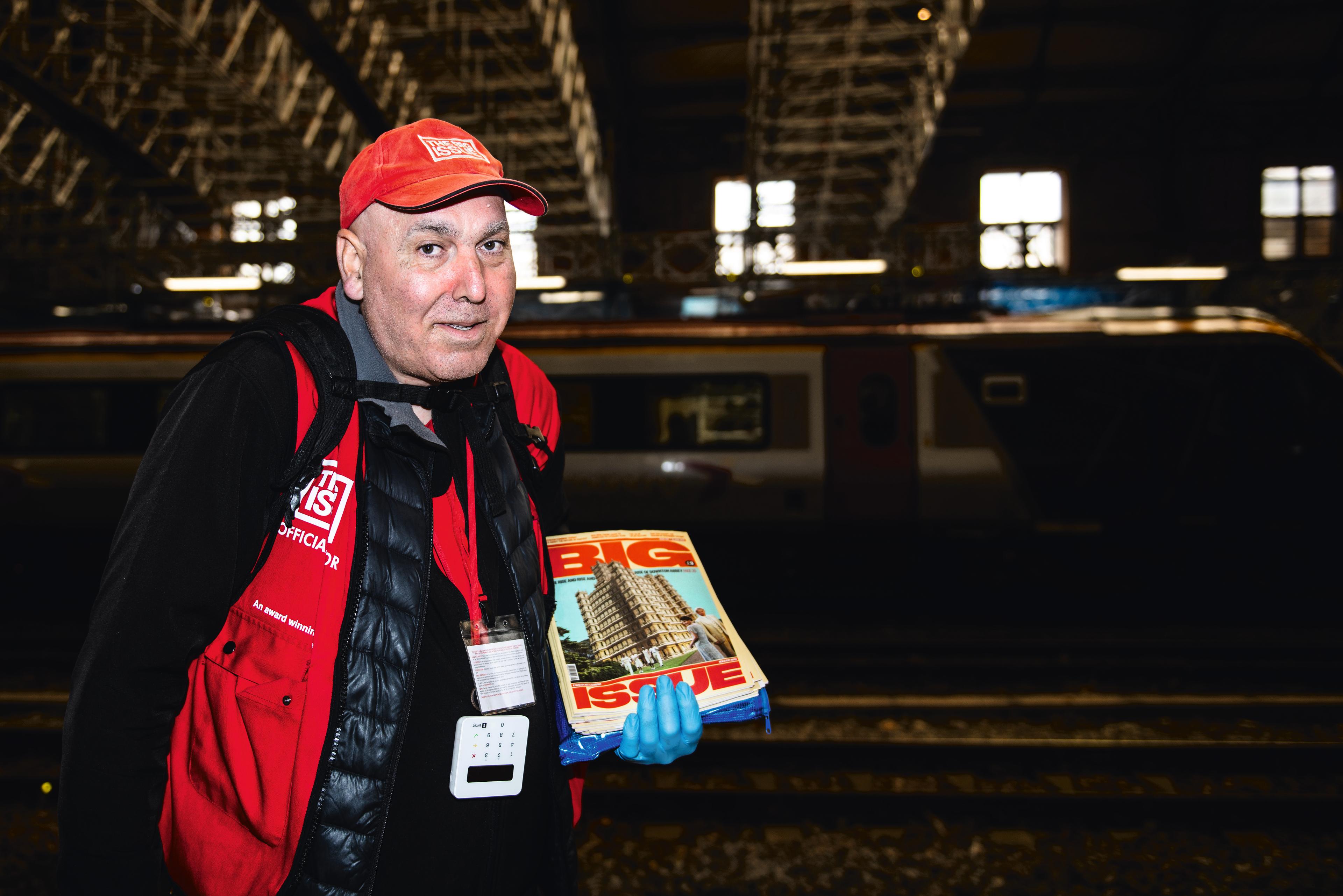 A man in red hat and vest holds magazines at a train station