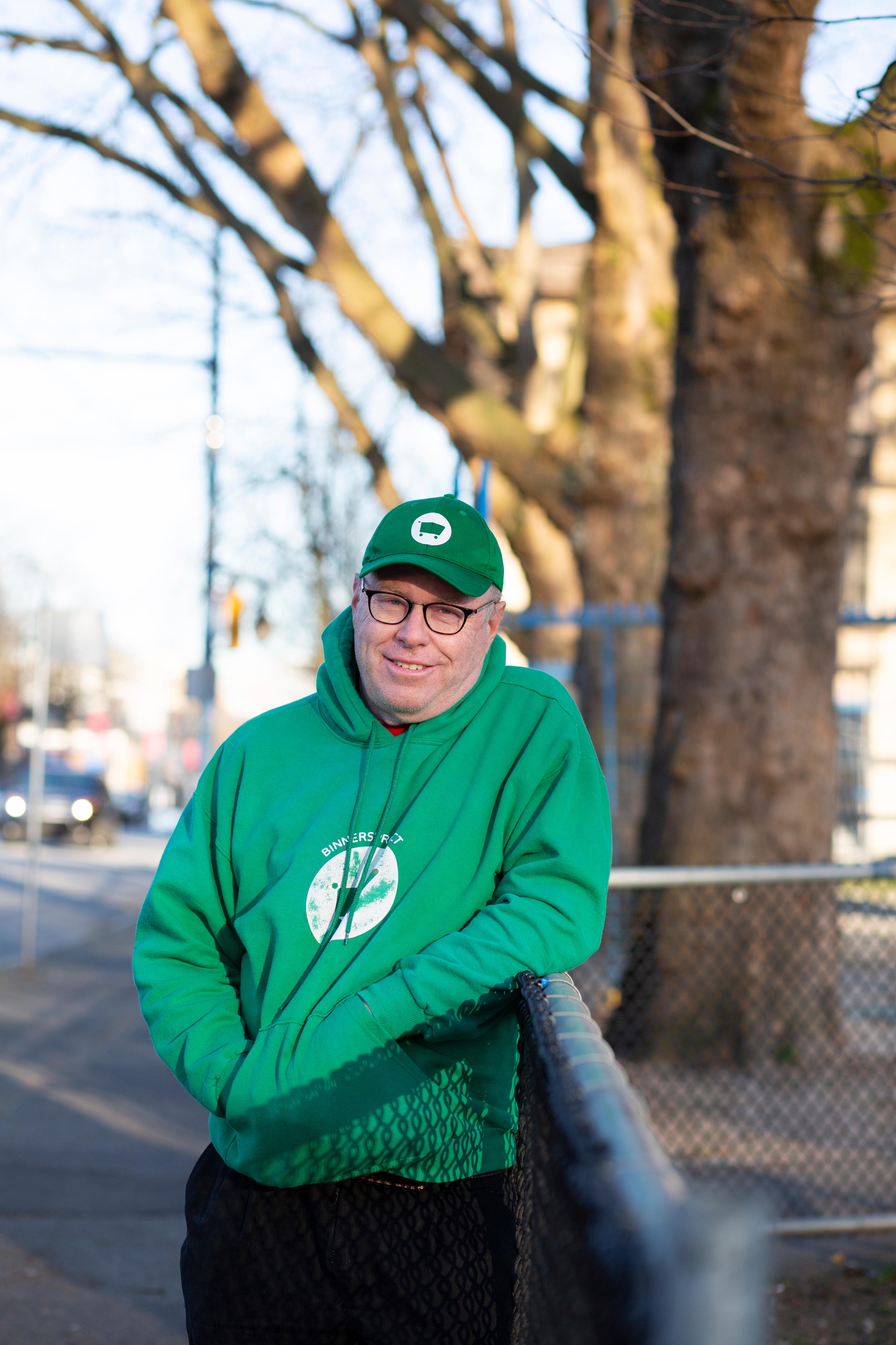 Mike McNeely leans against a fence wearing a green hat and hoodie