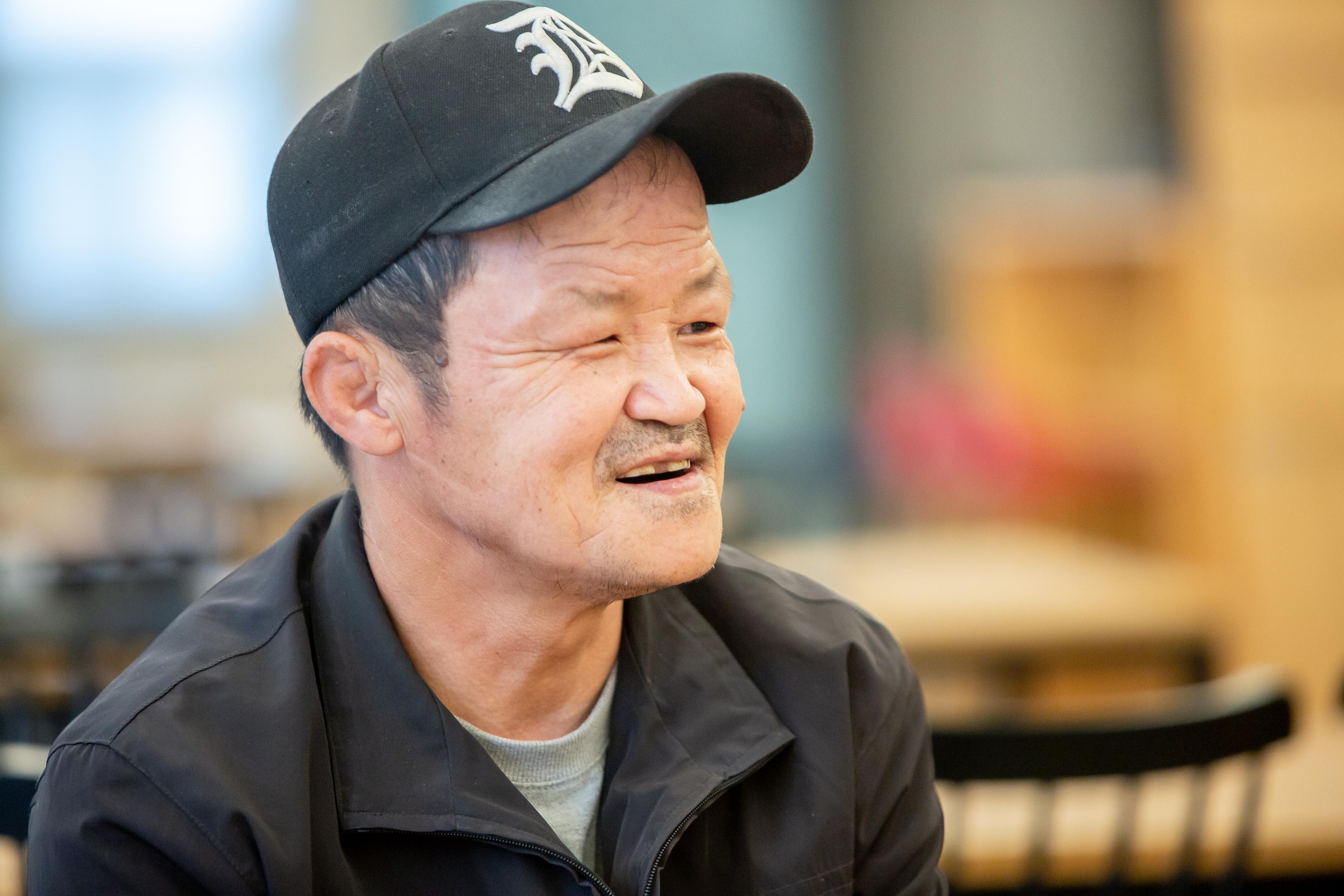 Big Issue Korea vendor Kwak Changgab is photographed in profile. He is smilign and wearing a black cap.