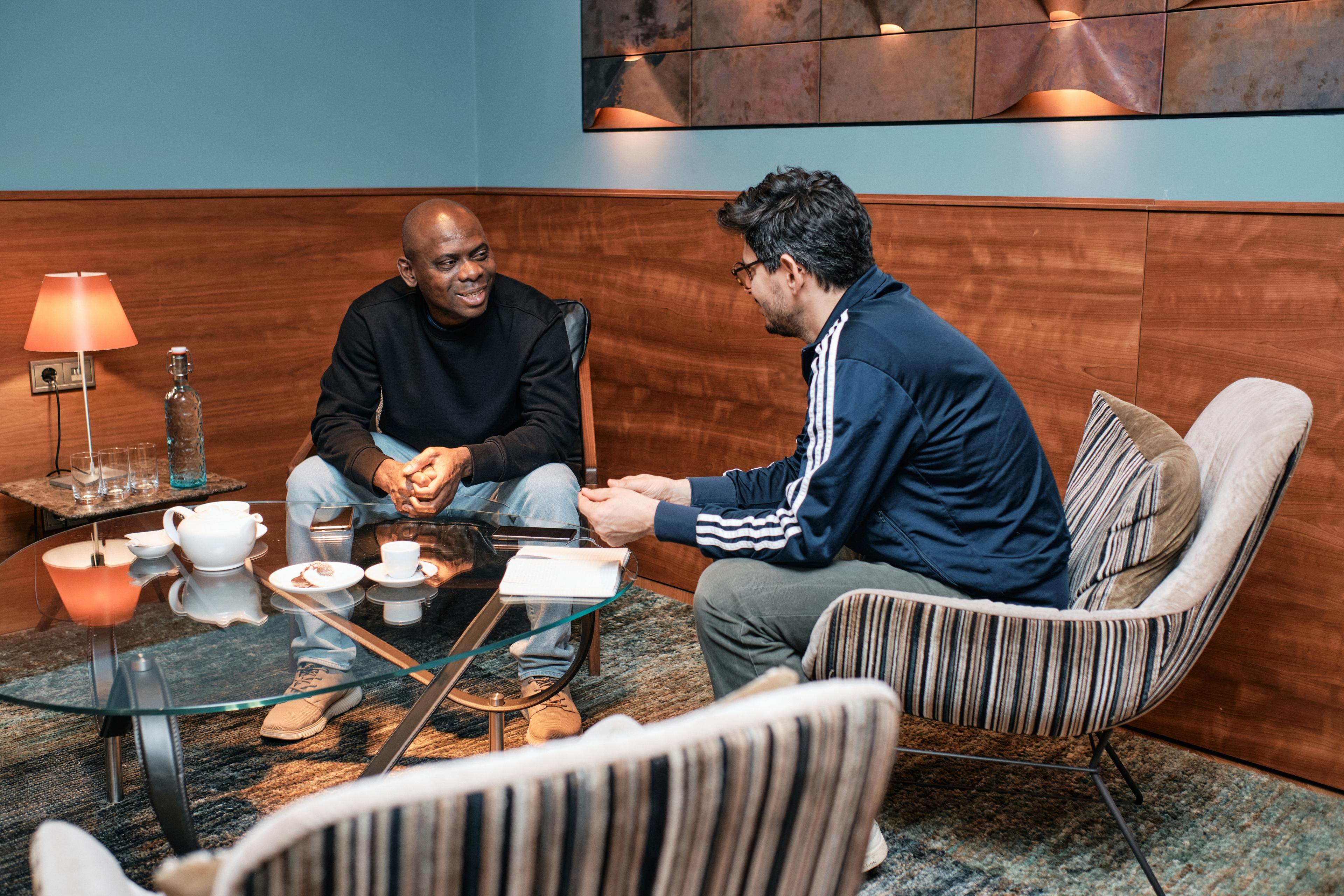 two men sitting in armchairs at a table, drinking coffee and chatting