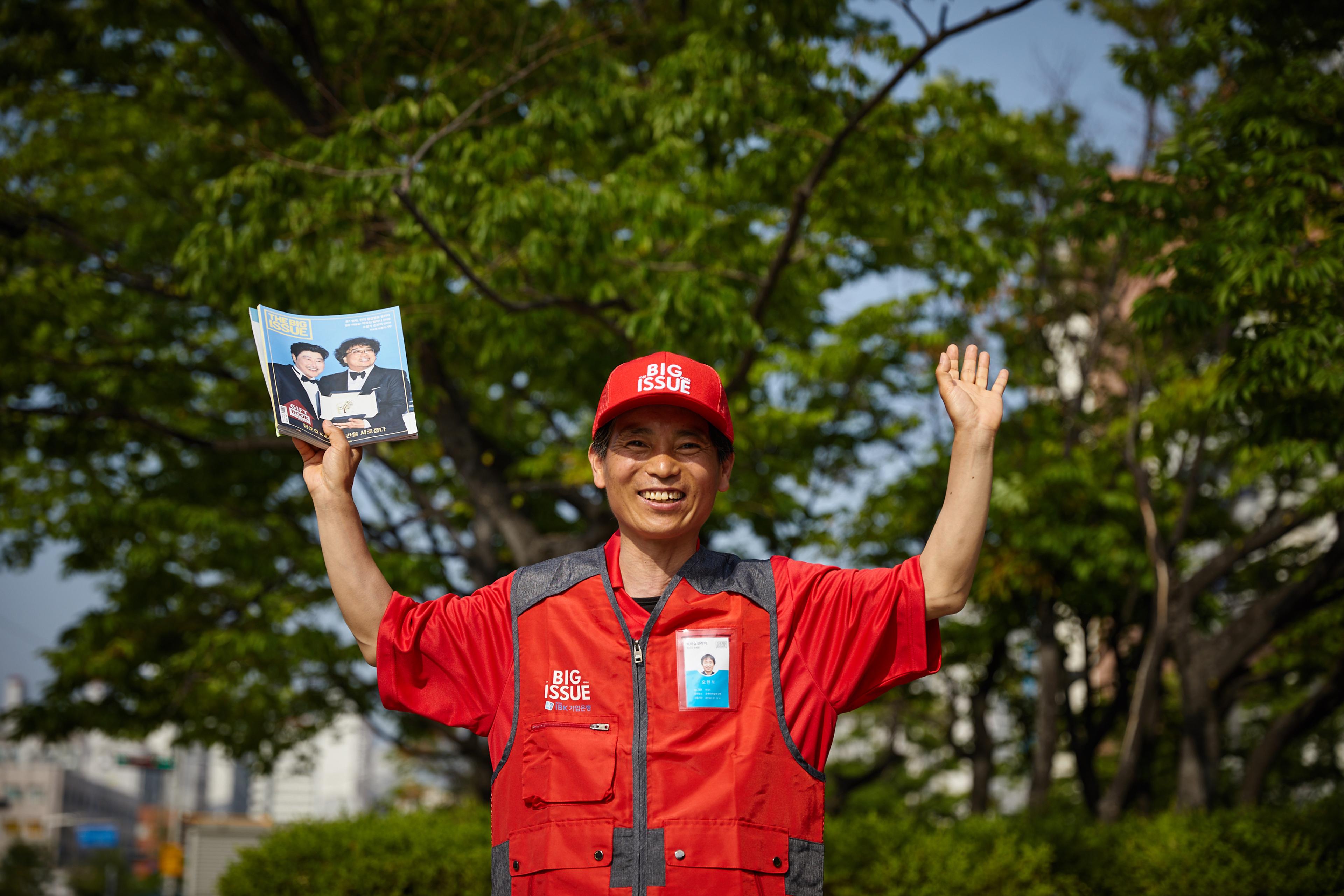 Big Issue Korea vendor Oh Hyun-suk, who has also been involved with the street soccer project, holds up a magazine wearing a red cap and red vest.