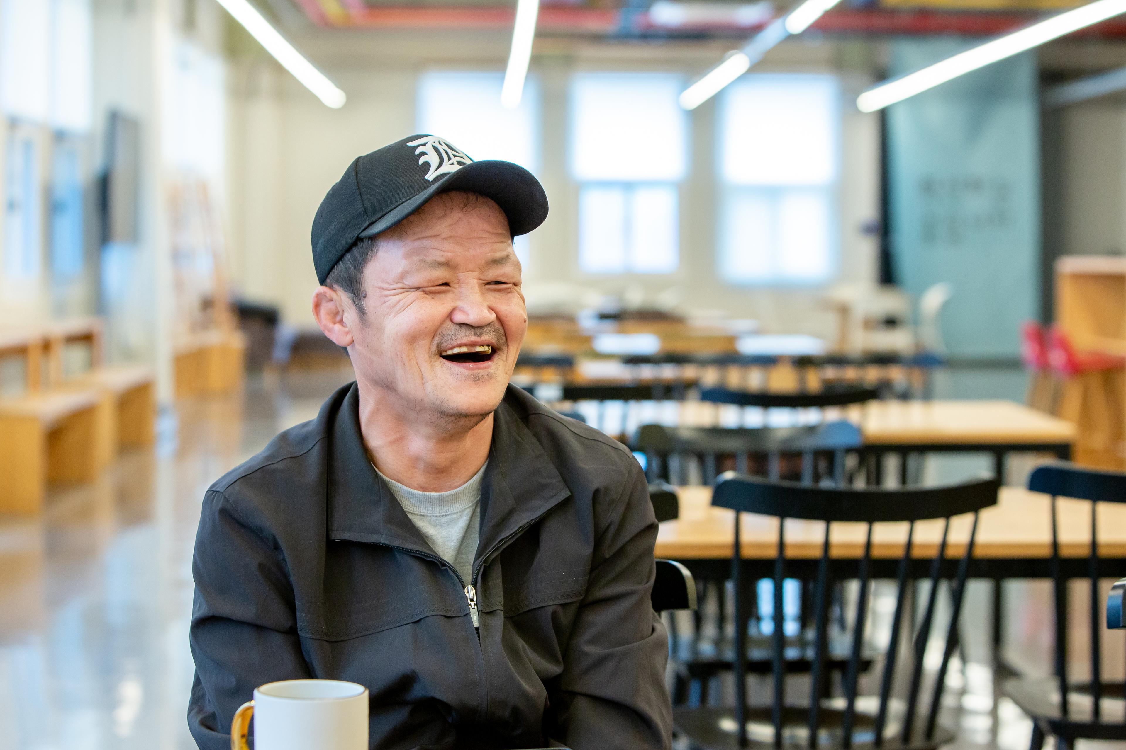 Big Issue Korea vendor Kwak Changgab is sitting in a cafe, laughing, and wearing a black cap and jacket.