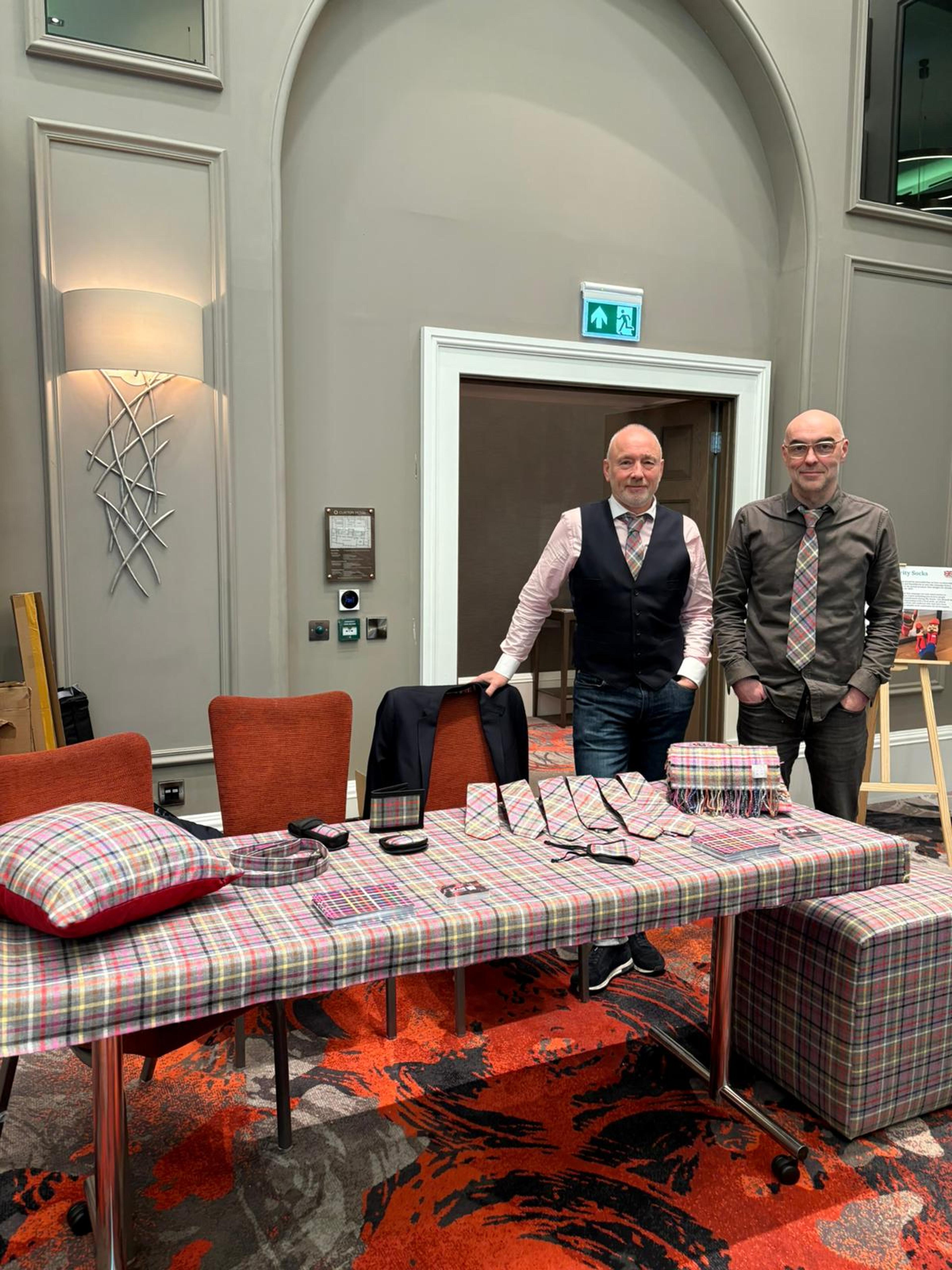 Brian (left) and Callum (right) of the Slanj Kilts team posing with a table of homeless tartan products