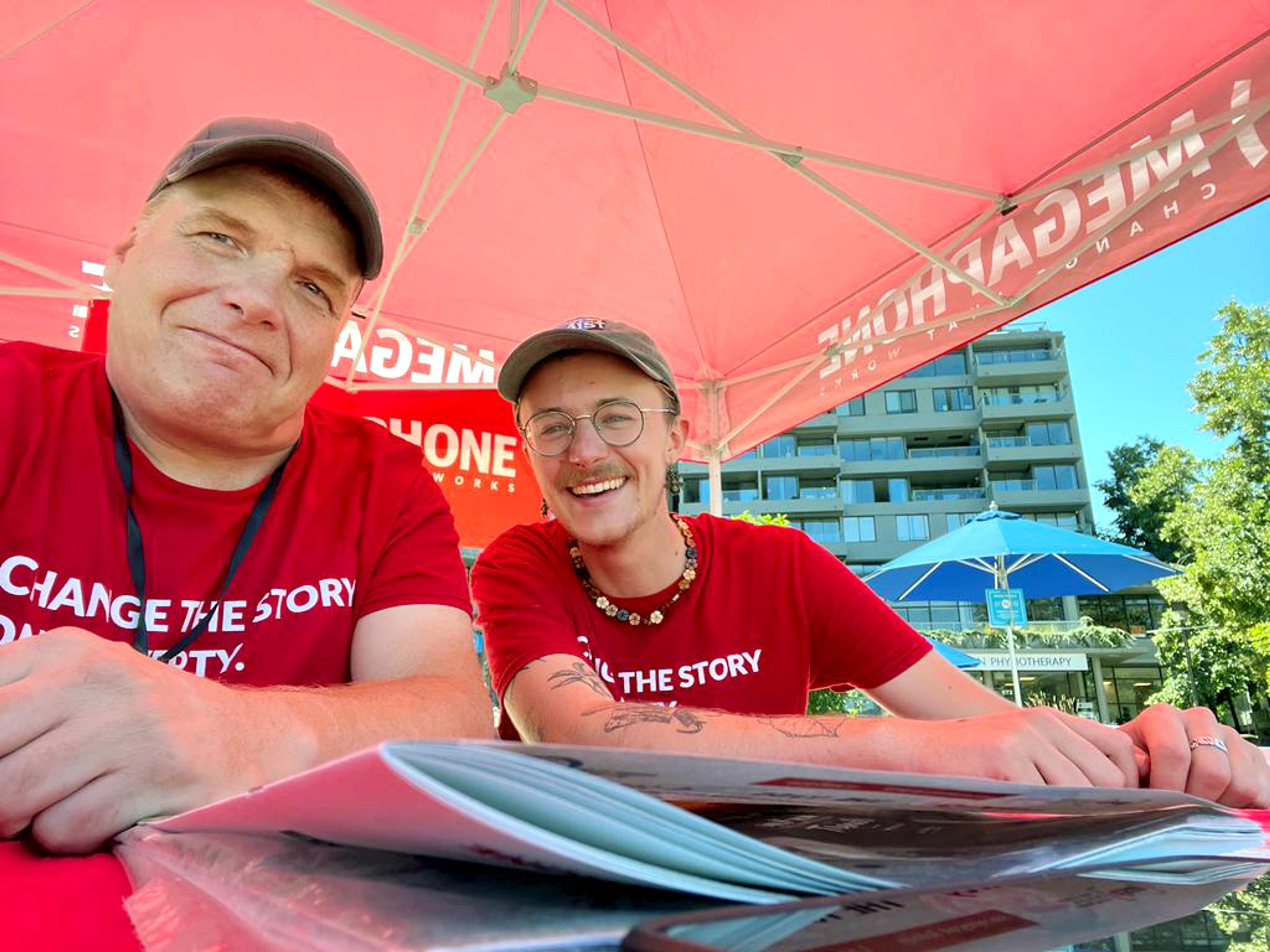 Two Megaphone street paper vendors smile at the camera wearing red t-shirts and caps