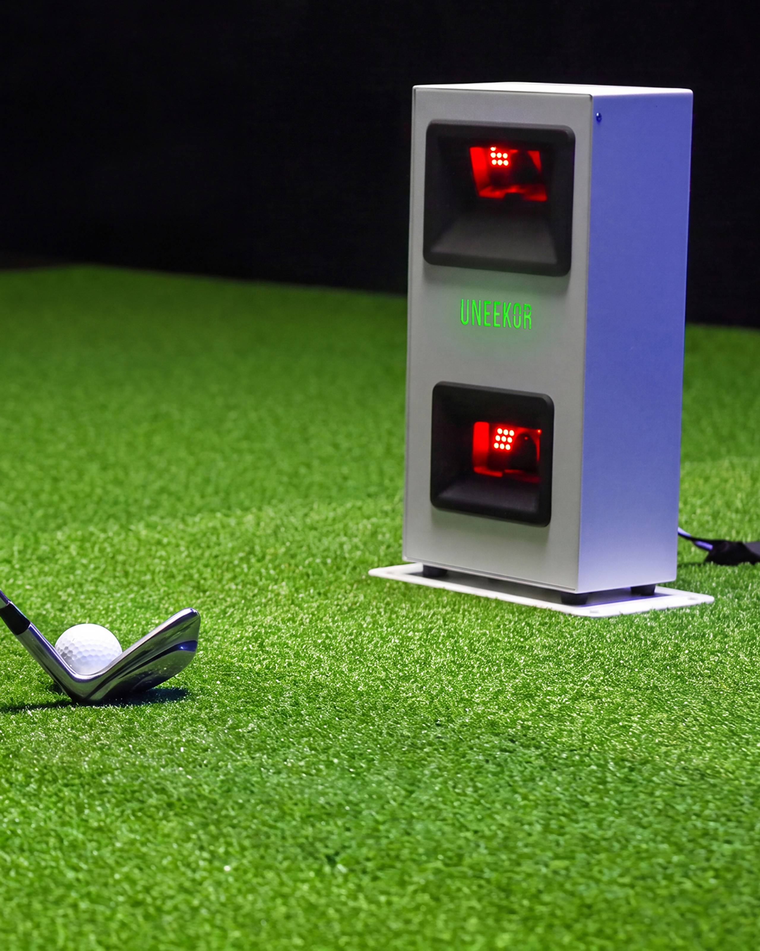 A golf club and ball on artificial turf, with a UNEKOR golf launch monitor in the background.