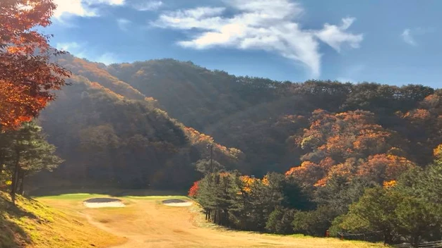 A golf course fairway and green surrounded by hills ablaze with autumn foliage under a blue sky.
