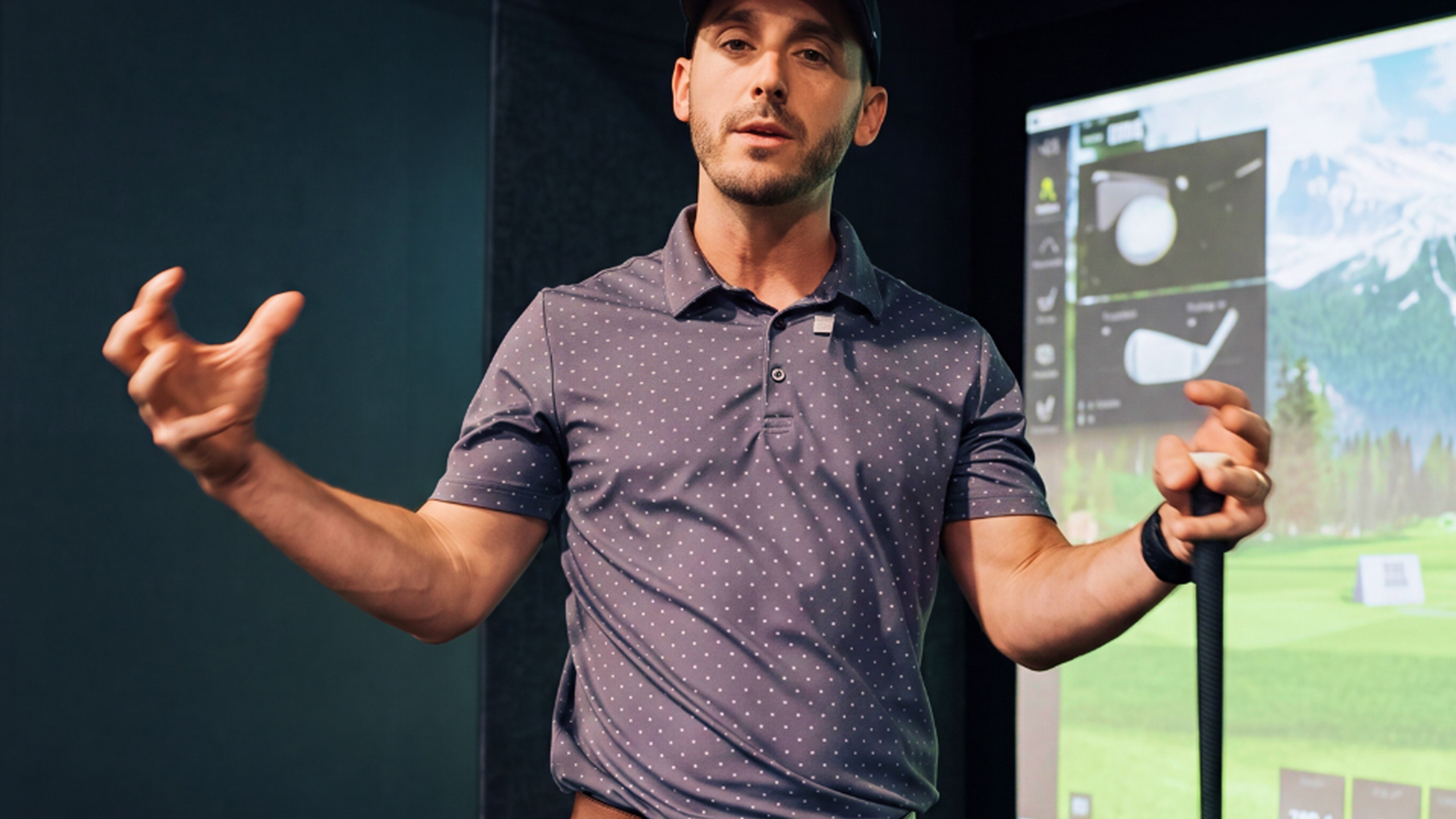 A man in a golf simulator, wearing a cap and polo shirt, holds a golf club and gestures, with a virtual golf course displayed behind him.