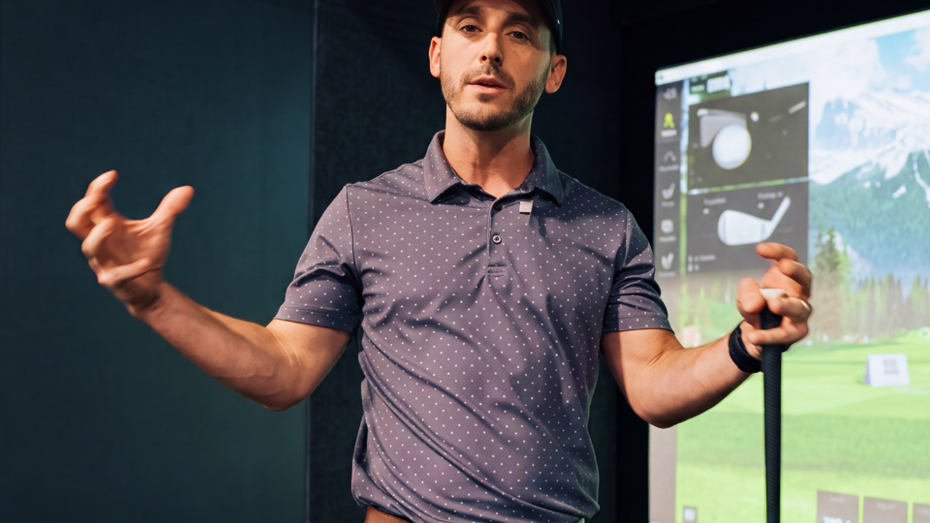 A man in a golf simulator, wearing a cap and polo shirt, holds a golf club and gestures, with a virtual golf course displayed behind him.
