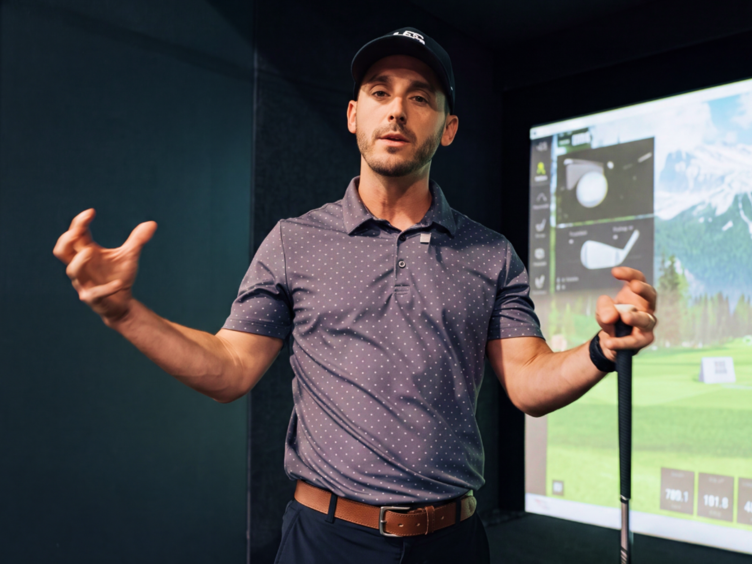 A man in a cap and polo shirt holds a golf club and gestures in front of a golf simulator screen.