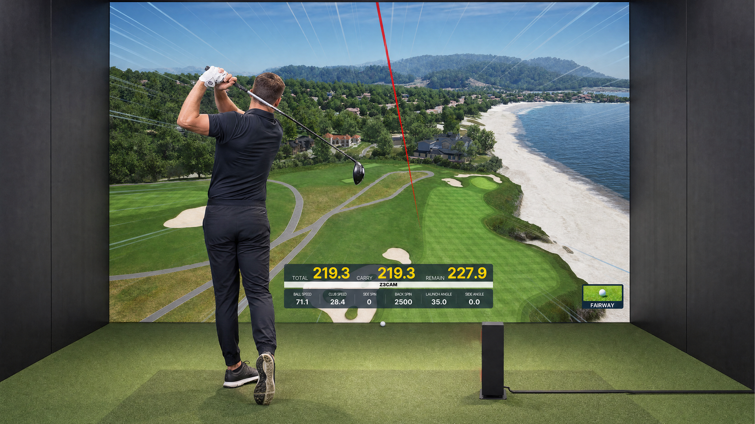 A man swings a golf club in a Uneekor indoor golf simulator, with a large screen displaying a virtual golf course by a beach and golf data.
