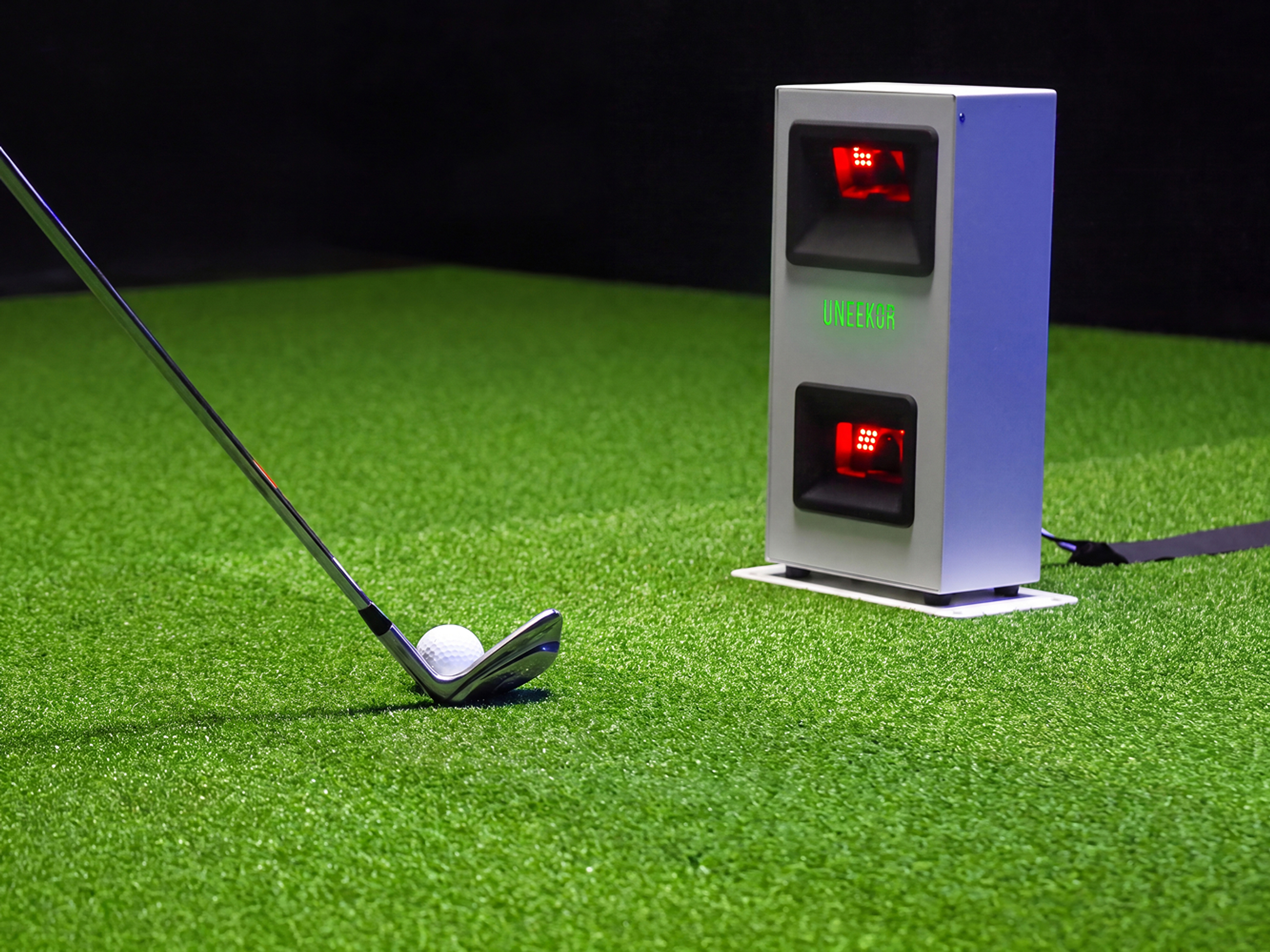 A golf club and ball on artificial green turf, next to a white golf launch monitor.