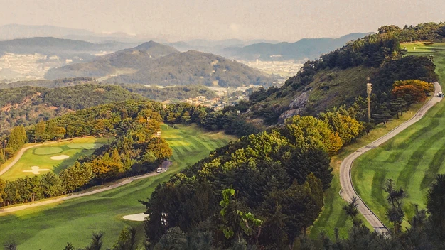 A golf course on forested hills, overlooking a town and mountains.