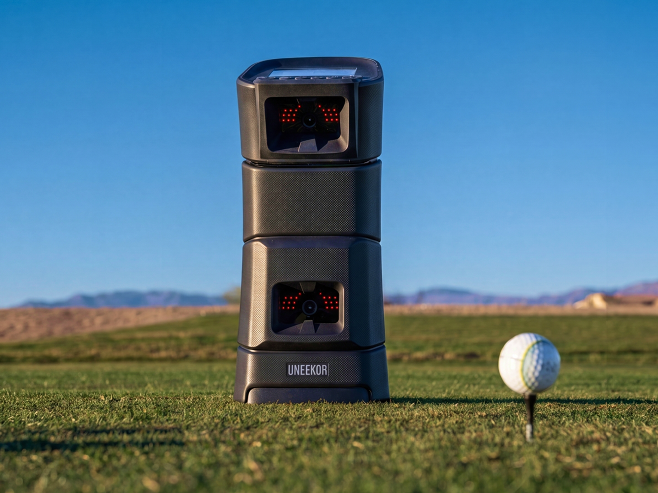 A black UNEEKOR golf launch monitor stands on a golf course next to a golf ball on a tee, under a clear blue sky.