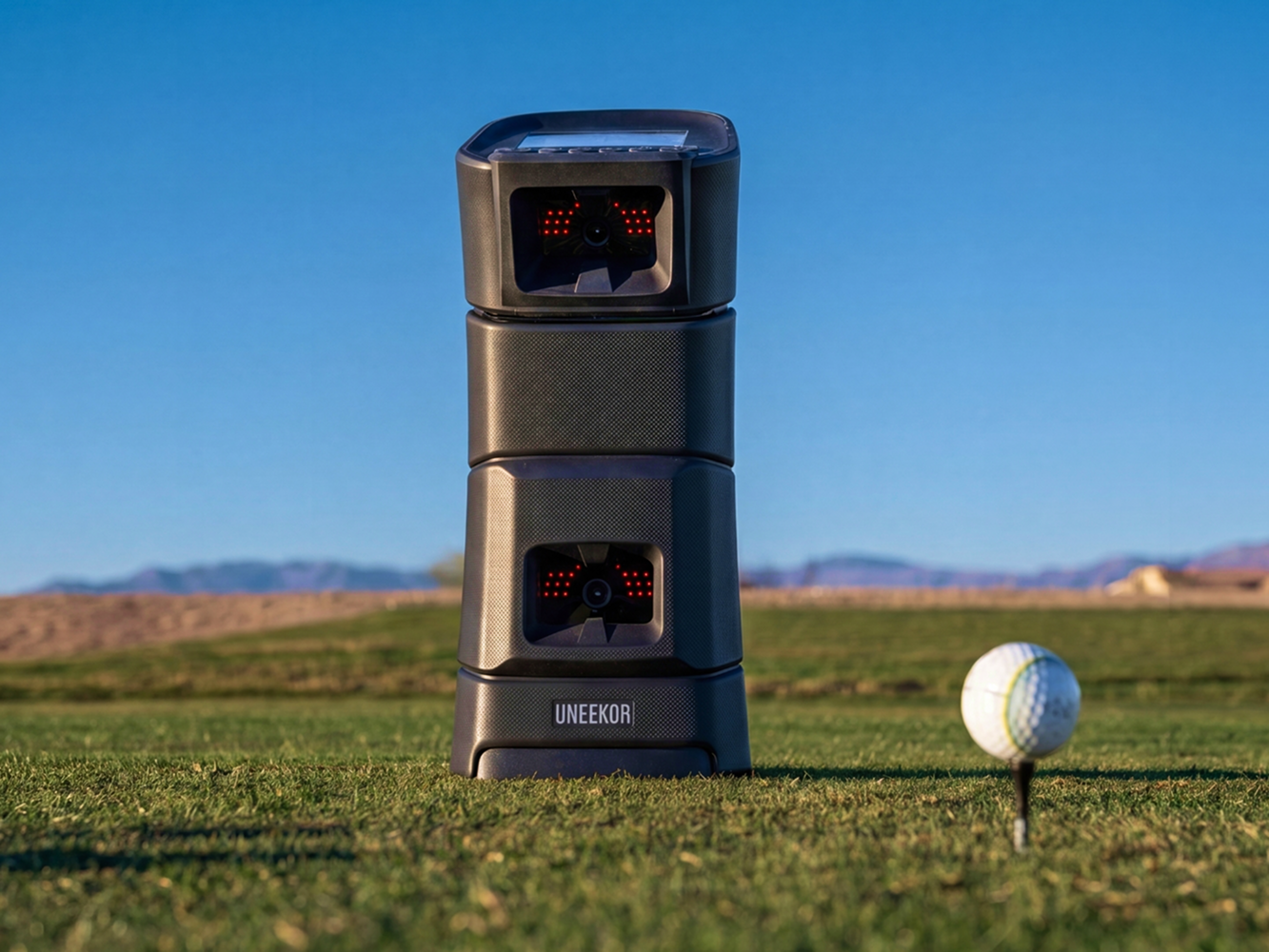 A black UNEEKOR golf launch monitor stands on a golf course next to a golf ball on a tee, under a clear blue sky.